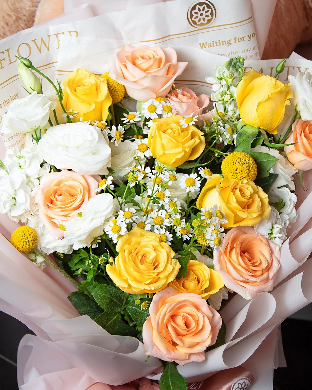A bouquet of mixed flowers including yellow roses, peach roses, white lisianthus, daisies, and yellow billy buttons, wrapped in decorative paper.