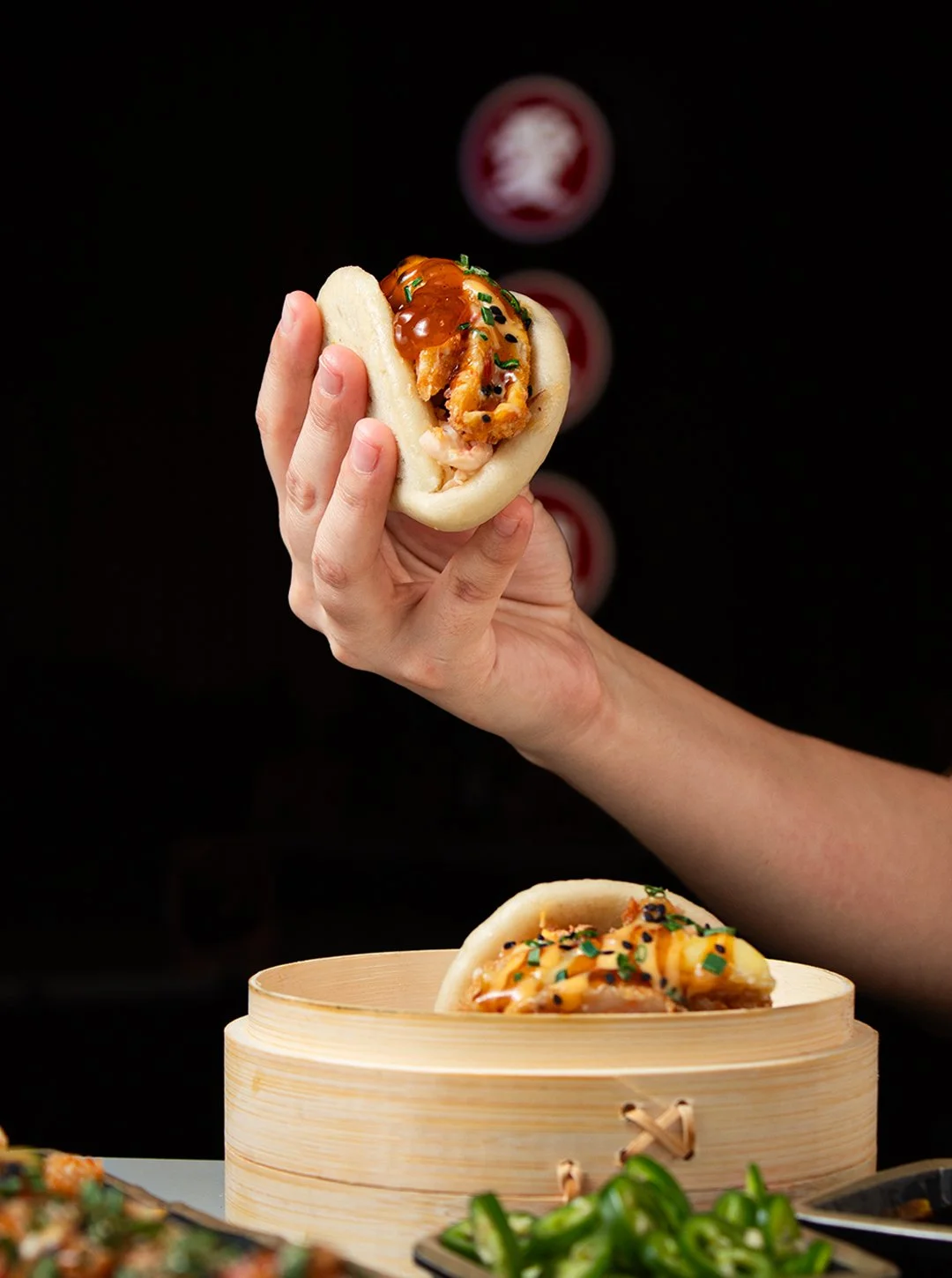 A hand holding a steamed bun filled with a cooked lobster tail and topped with sauce and chopped green onions, with a bamboo steamer and other food items visible in the background.