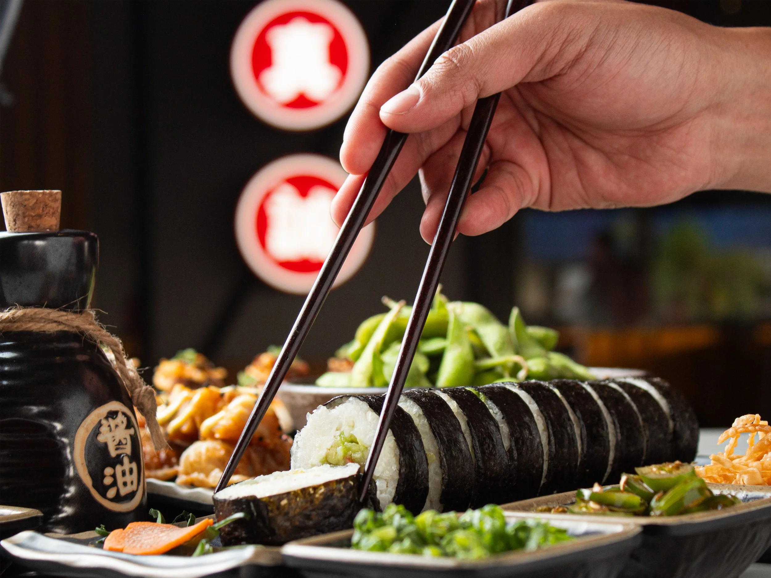 Hand using chopsticks picking up a piece of sushi, with various sushi and side dishes on a table.