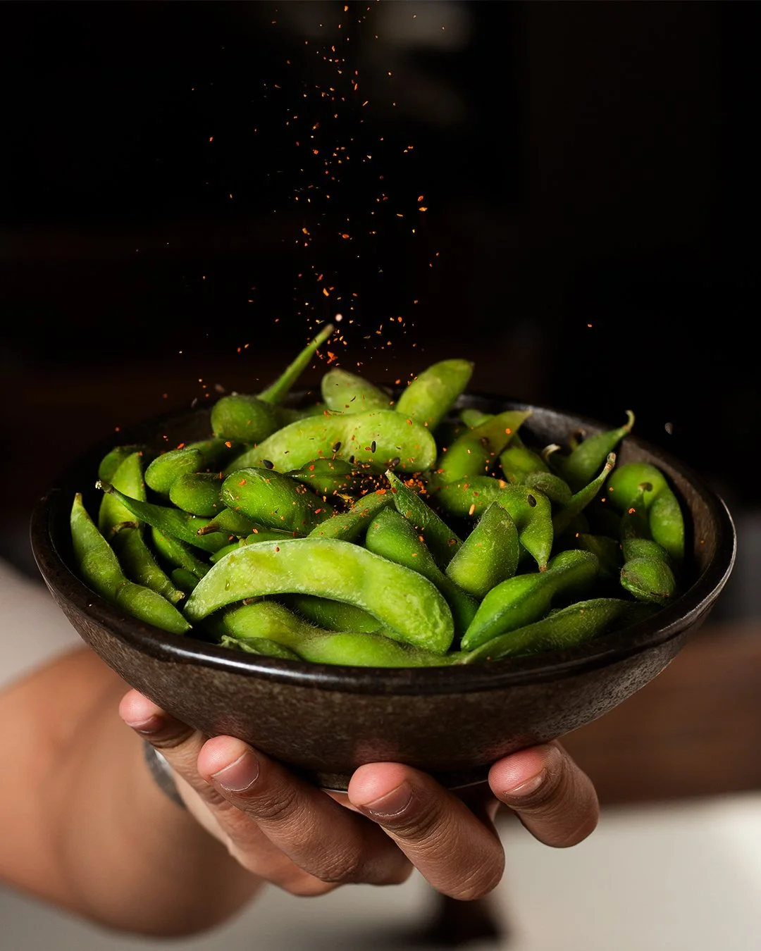 A hand holding a black bowl filled with green edamame beans, with red pepper flakes being sprinkled over them.