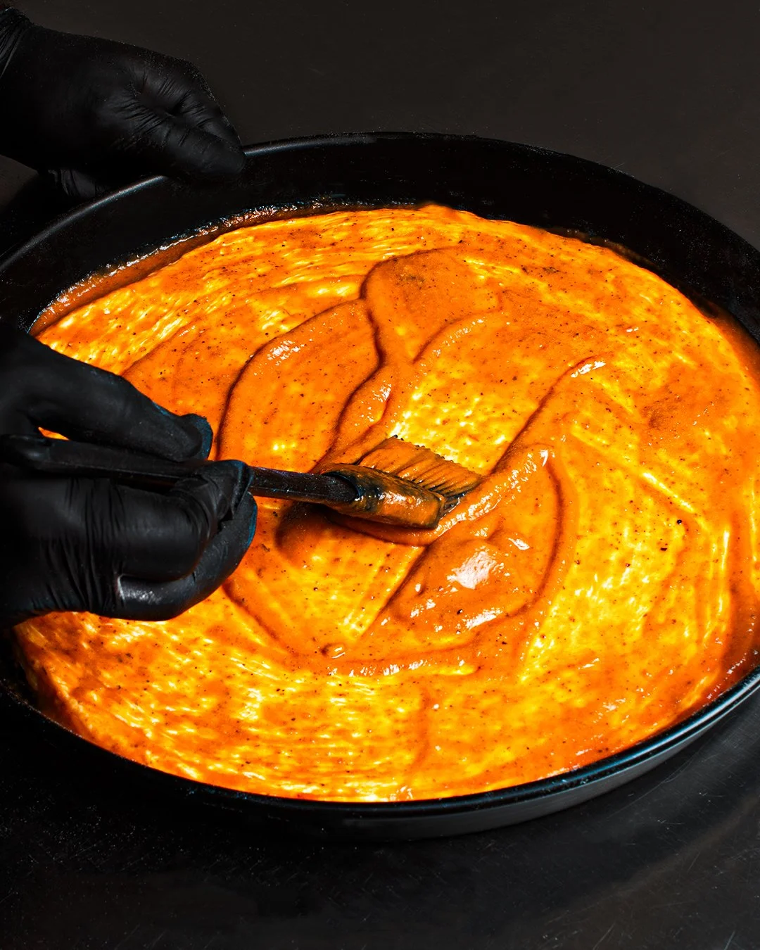 Person wearing black gloves spreading orange-colored sauce over flat fish fillets in a pan.