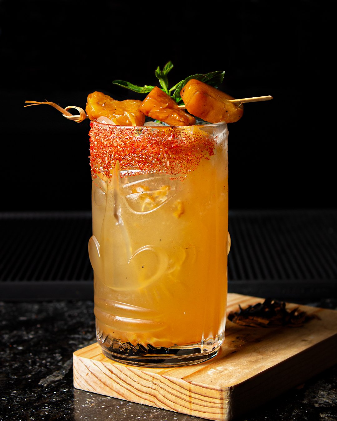 A cocktail glass with an orange-colored drink, garnished with grilled pineapple skewers and a sprig of mint, with chili powder rim, placed on a wooden coaster against a black background.