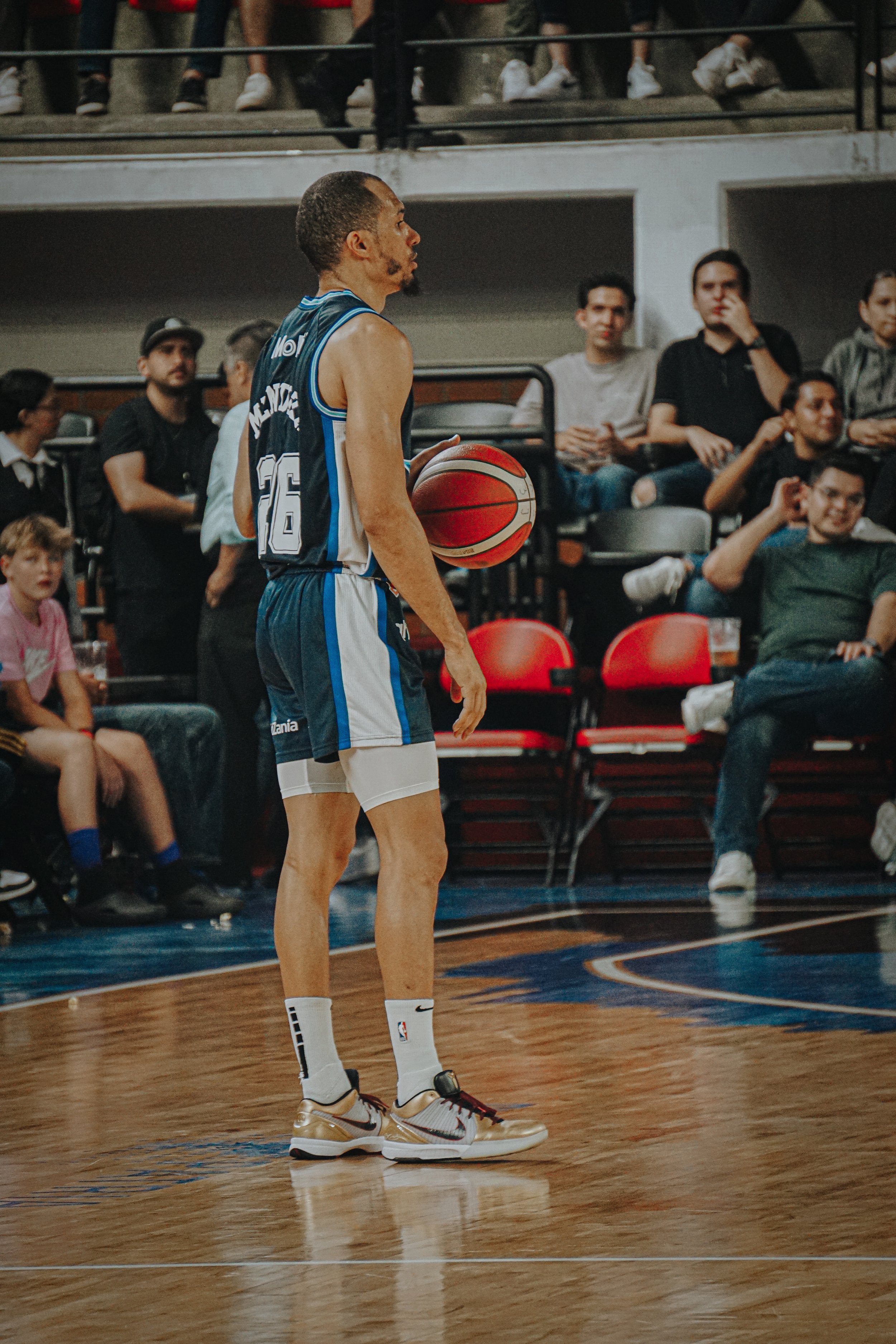 A basketball player standing on a court holding a basketball, with spectators seated in the background.