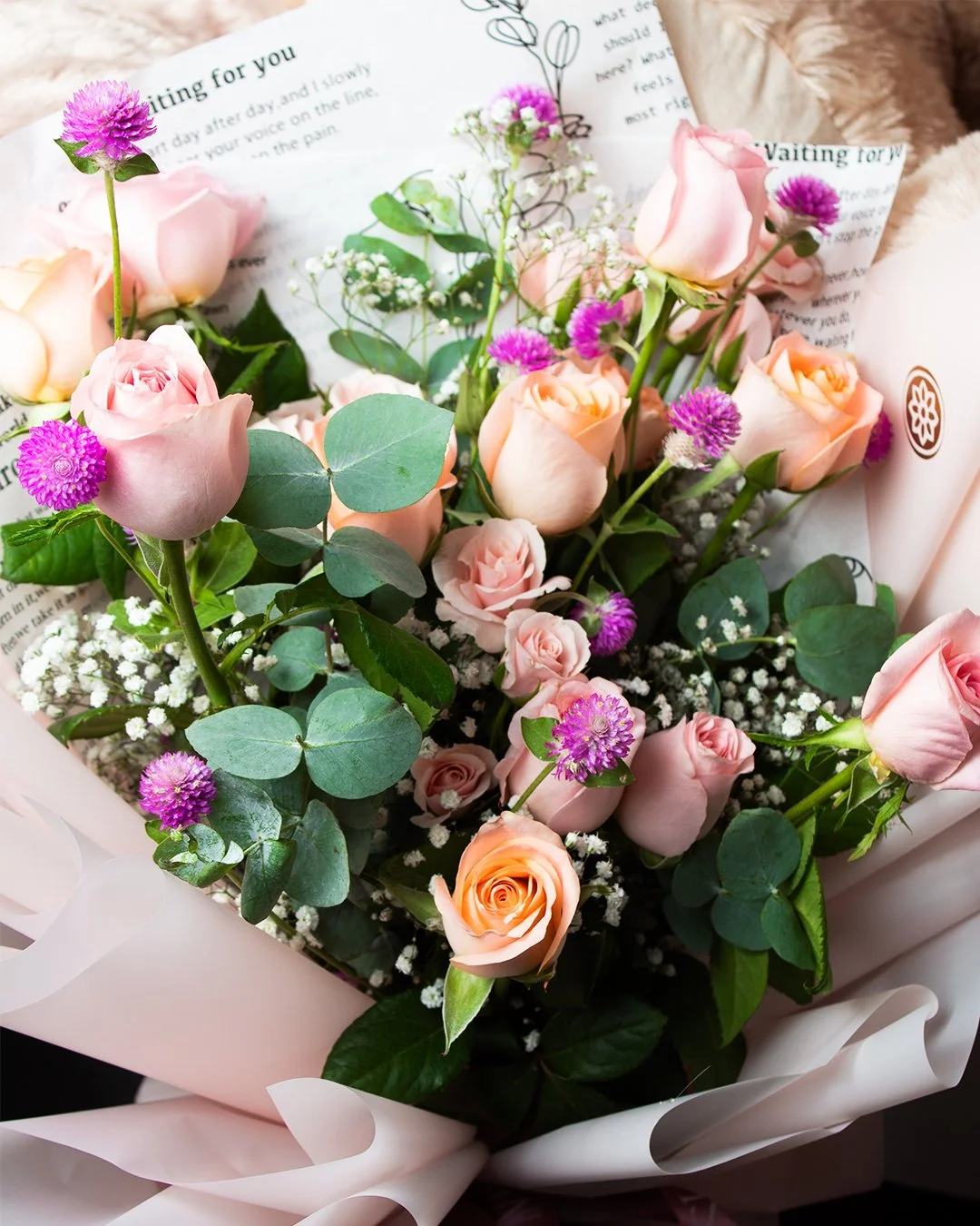 A bouquet of pink and peach roses, pink globe amaranth flowers, white baby's breath, and eucalyptus leaves wrapped in pink paper.