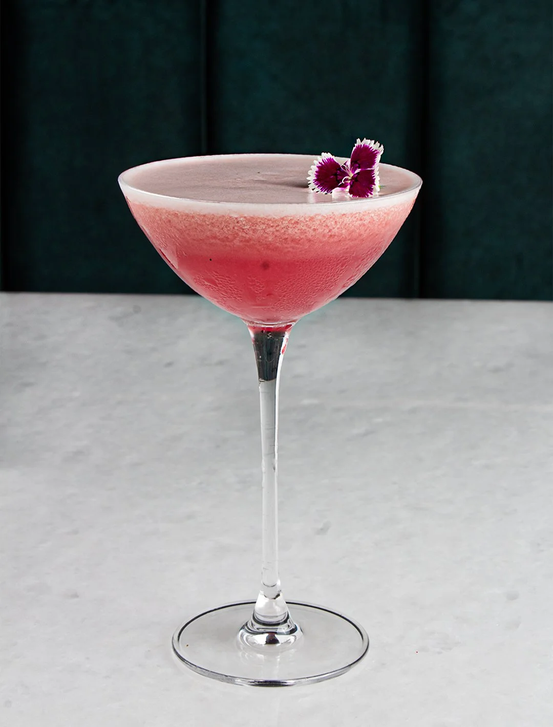 A pink cocktail in a coupe glass garnished with purple and white flowers, placed on a light gray surface with a dark background.