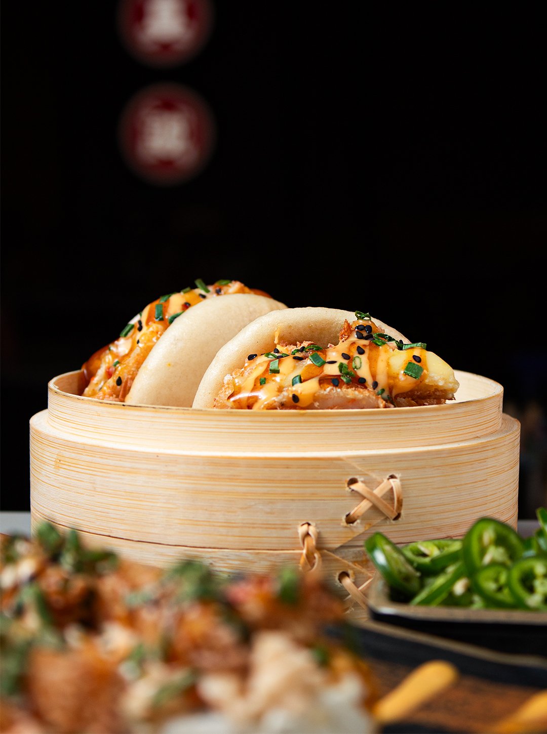 Two bao buns filled with fried chicken, drizzled with orange sauce, topped with chopped chives and black sesame seeds in a round bamboo steamer, with a dish of sliced green peppers in the foreground.