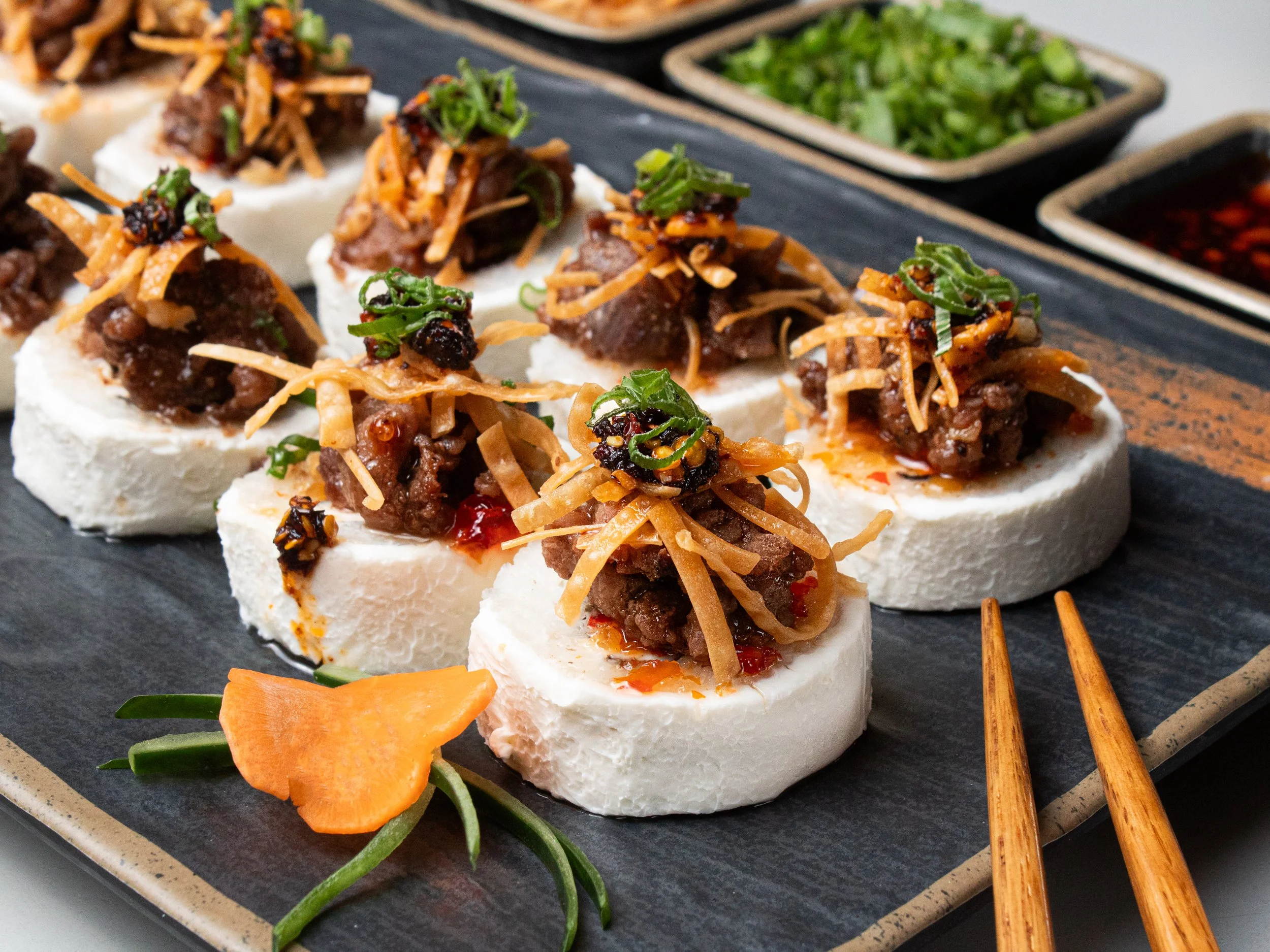 Chinese beef and shredded ginger served on rice cakes, garnished with chopped green onions on a black platter with dipping sauces and vegetables.
