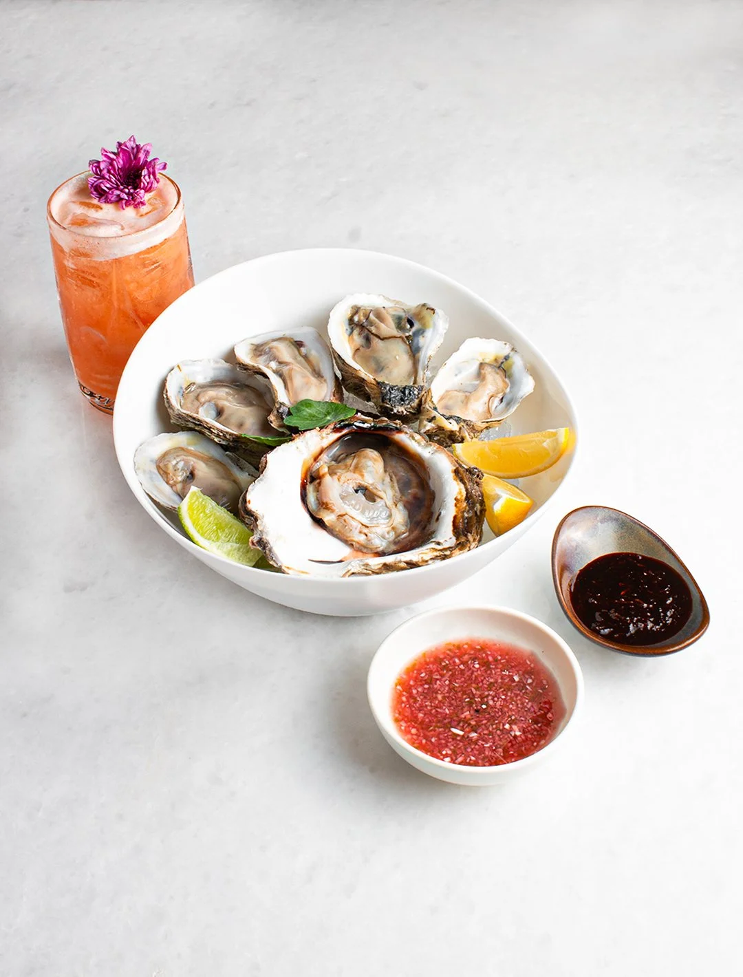 A white bowl filled with opened oysters on the half shell, garnished with lime and lemon wedges, placed on a light surface. To the right are small bowls of red and dark sauces, and a tall glass of pink cocktail with a purple flower on top.