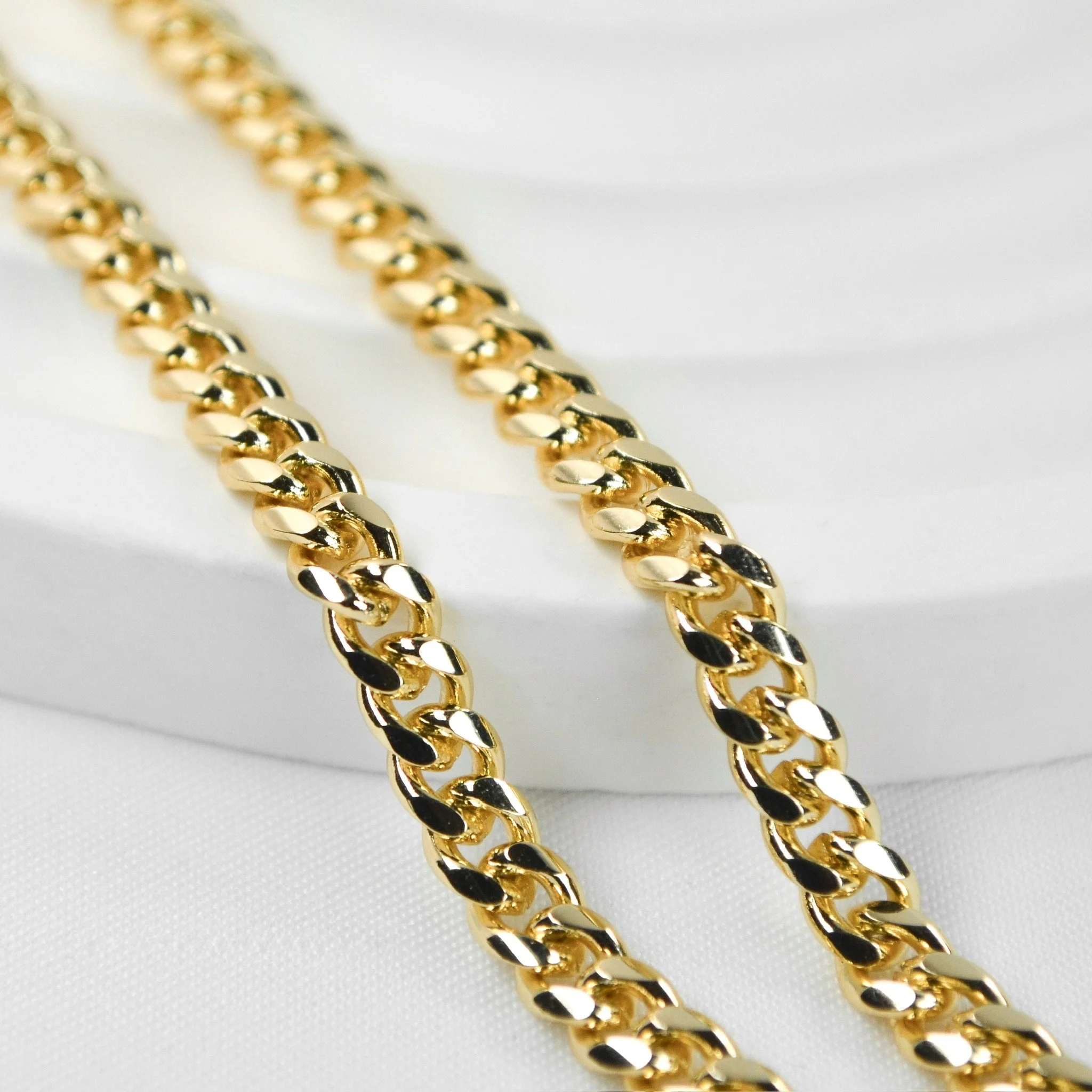 Close-up of two gold chain necklaces with a shiny, polished finish, resting on a white textured surface.