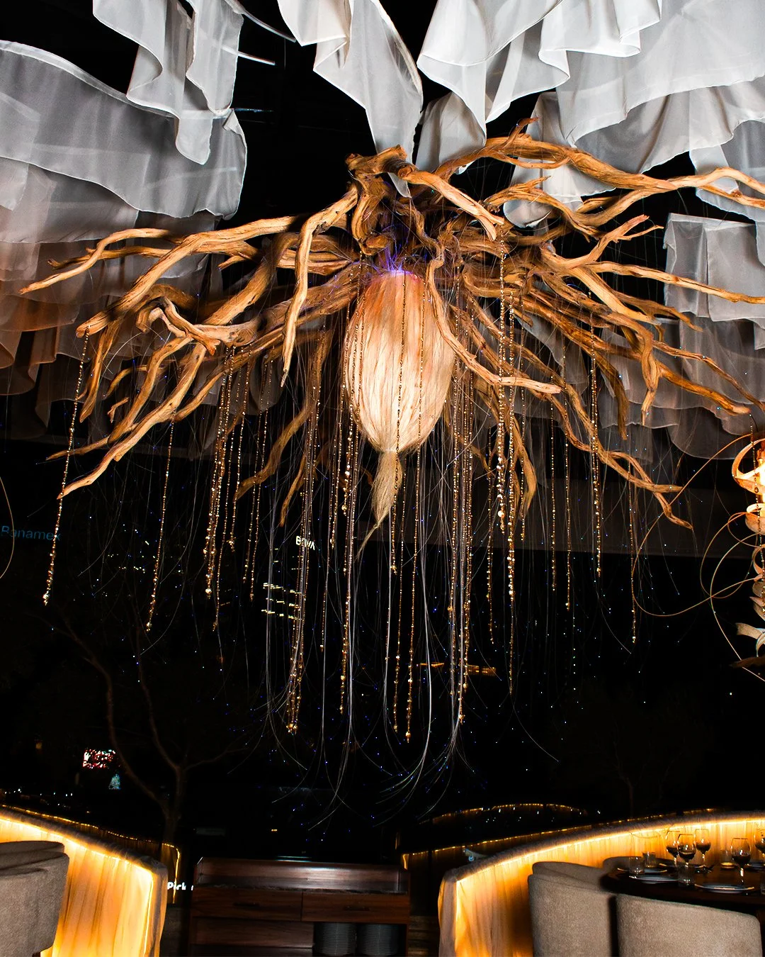 Decorative installation resembling a tree branch with a large light bulb in the center, hanging from the ceiling of a restaurant or event space, with white draped fabric and hanging string lights in the background.