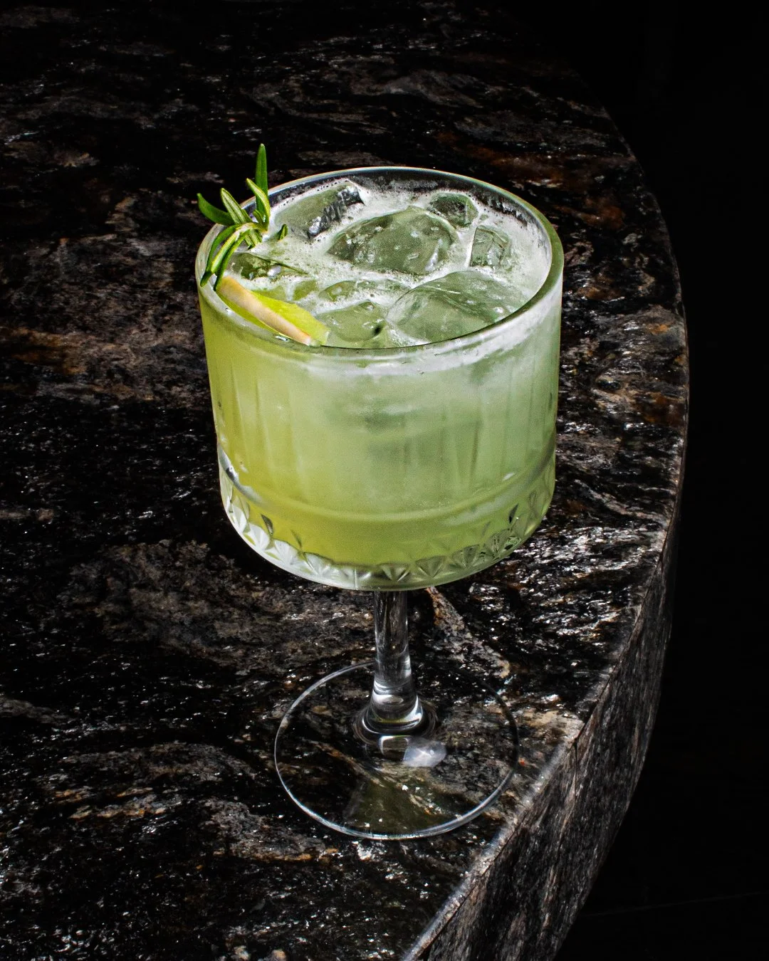 A green cocktail with ice cubes in a short, ornate glass garnished with a sprig of rosemary and a lemon wedge, on a dark marble surface.