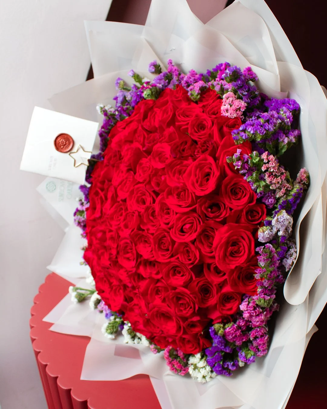 A large bouquet of red roses surrounded by purple and pink flowers, wrapped in white paper.
