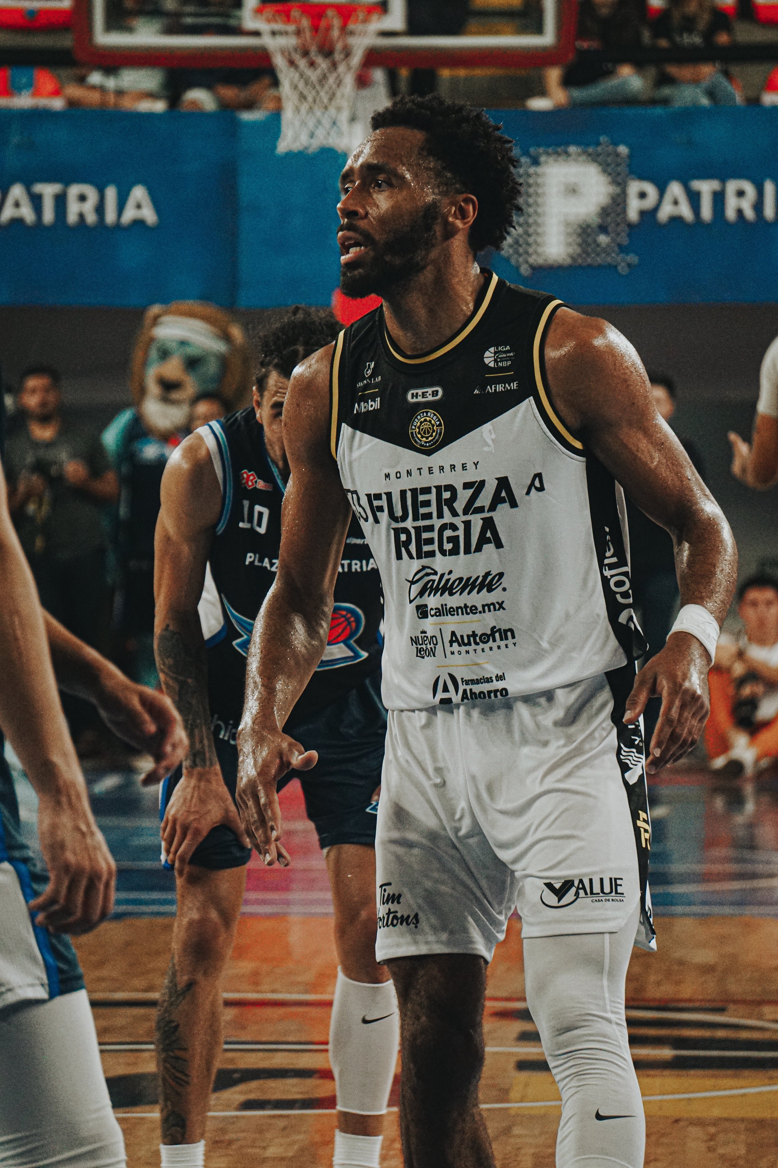 A basketball player on the court during a game, wearing a black and white jersey with logos, sweat on his arm, with other players and a crowd in the background.