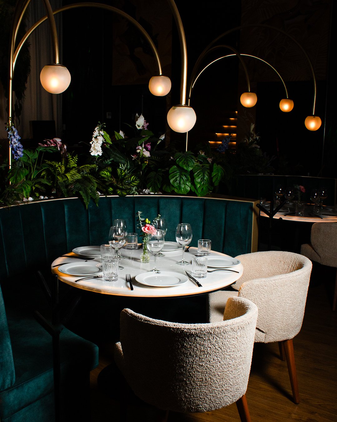 A dining table set for six with white plates, glasses, and cutlery in a dimly lit restaurant. The table has a small pink flower centerpiece. Surrounding the table are plush beige chairs and a green curved banquette. Overhead, curved gold lamps with r