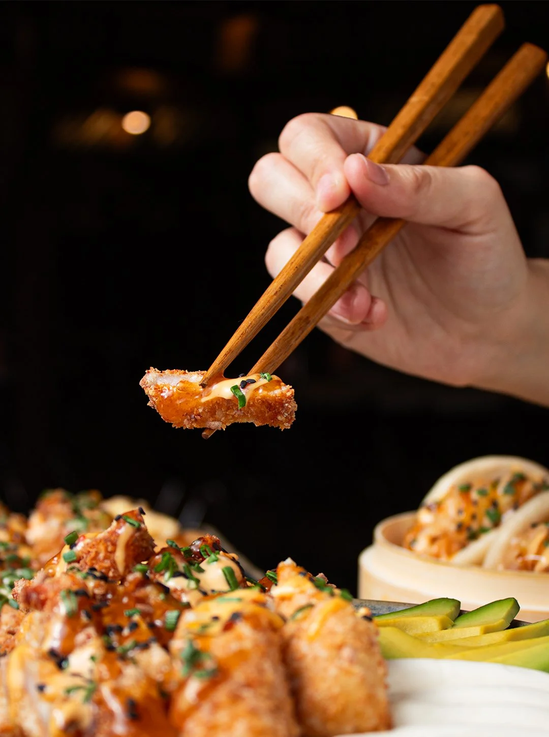Person holding chopsticks picking up a piece of fried sushi topped with sauce and green onions, with other sushi rolls, sliced cucumbers, and noodle dishes in the background.