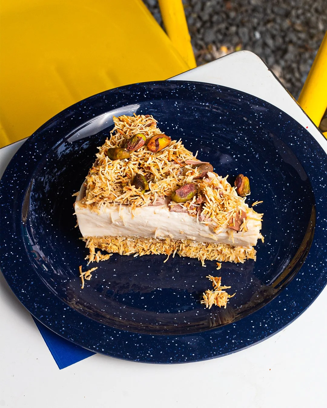 Slice of cream cheese cake topped with shredded coconut and pistachios on a dark blue speckled plate.