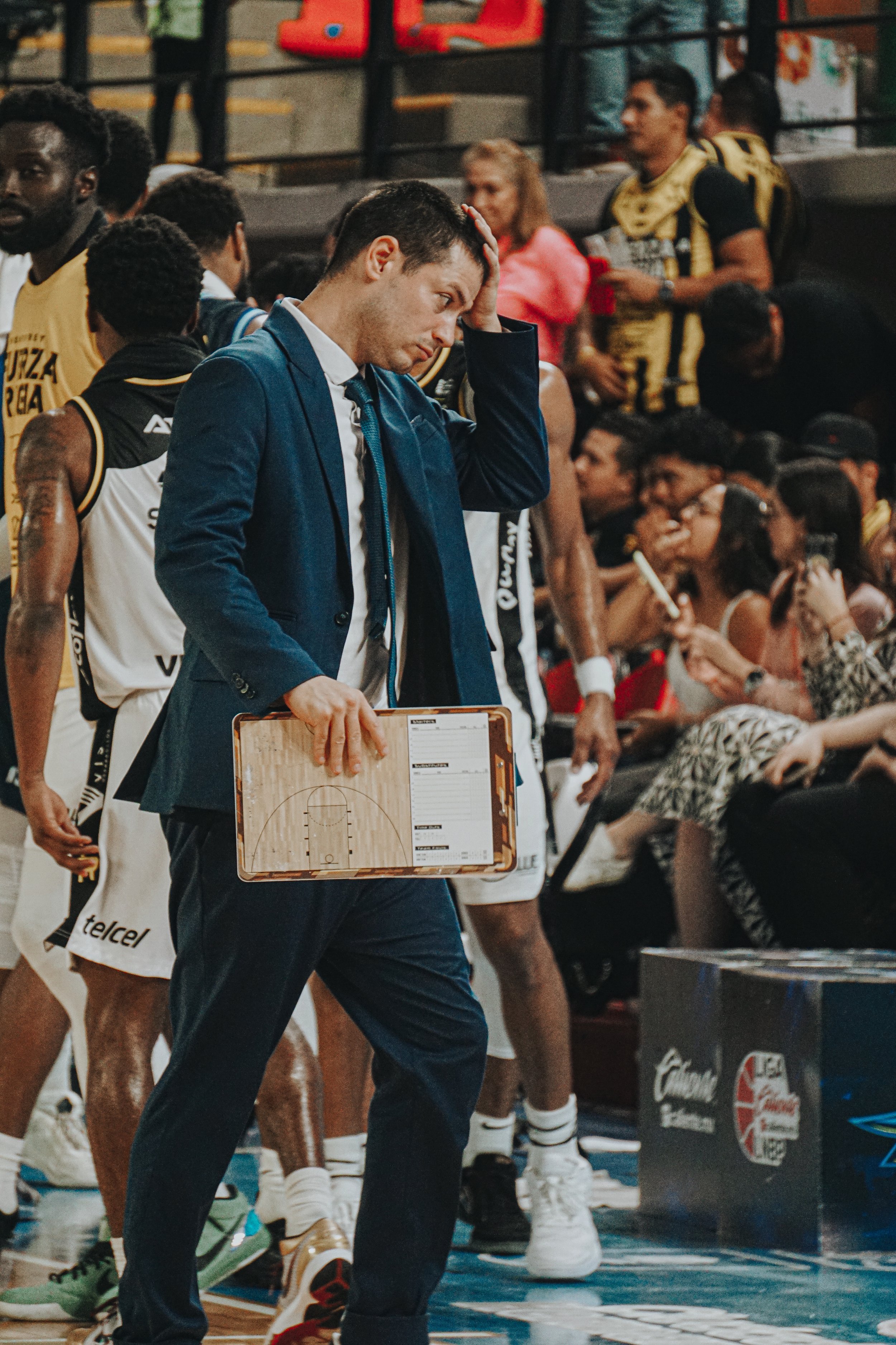 Basketball coach holding a playboard on the sideline of a game, with players and spectators in the background