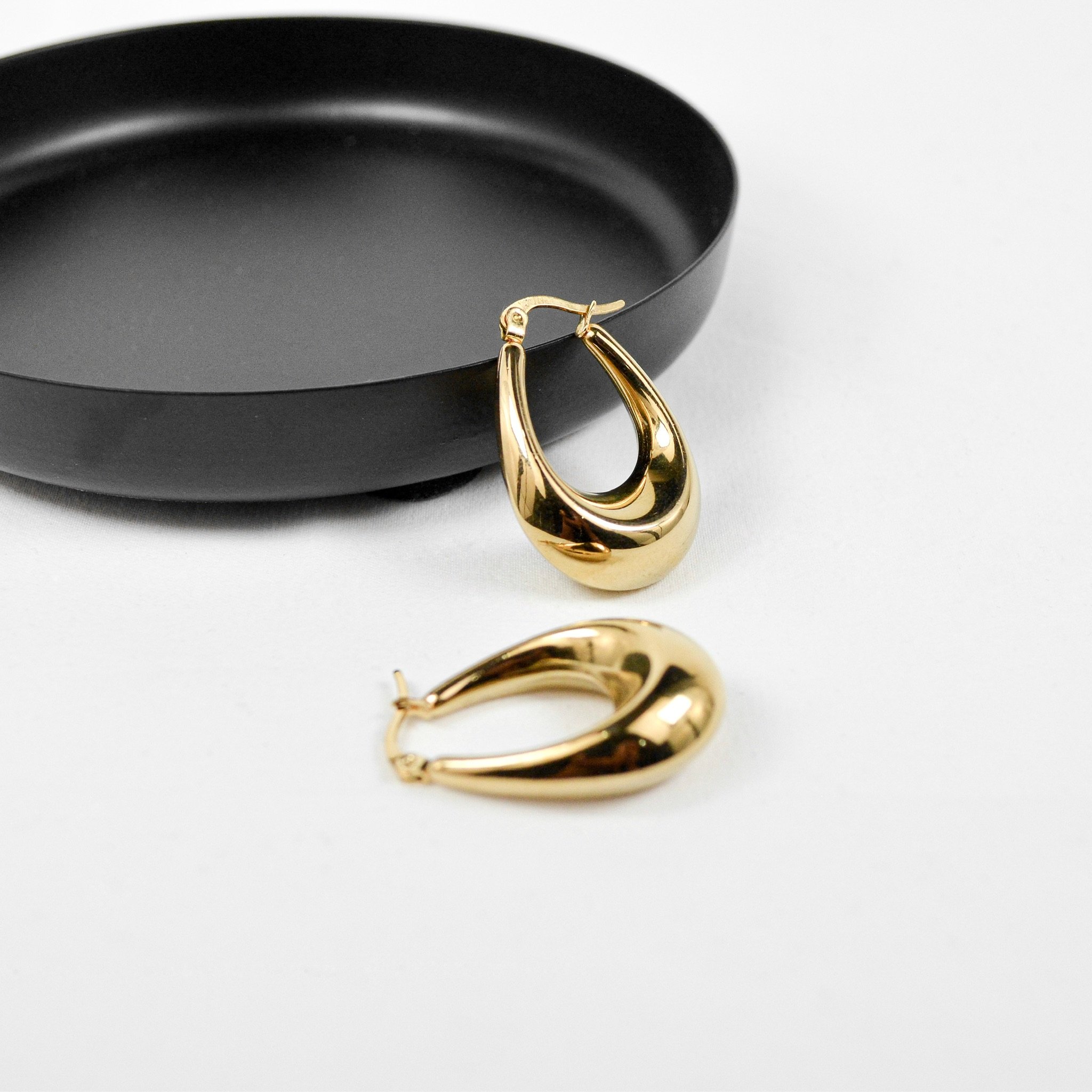 Gold hoop earrings with a thick, teardrop-shaped design, placed on a white surface with a black round dish.