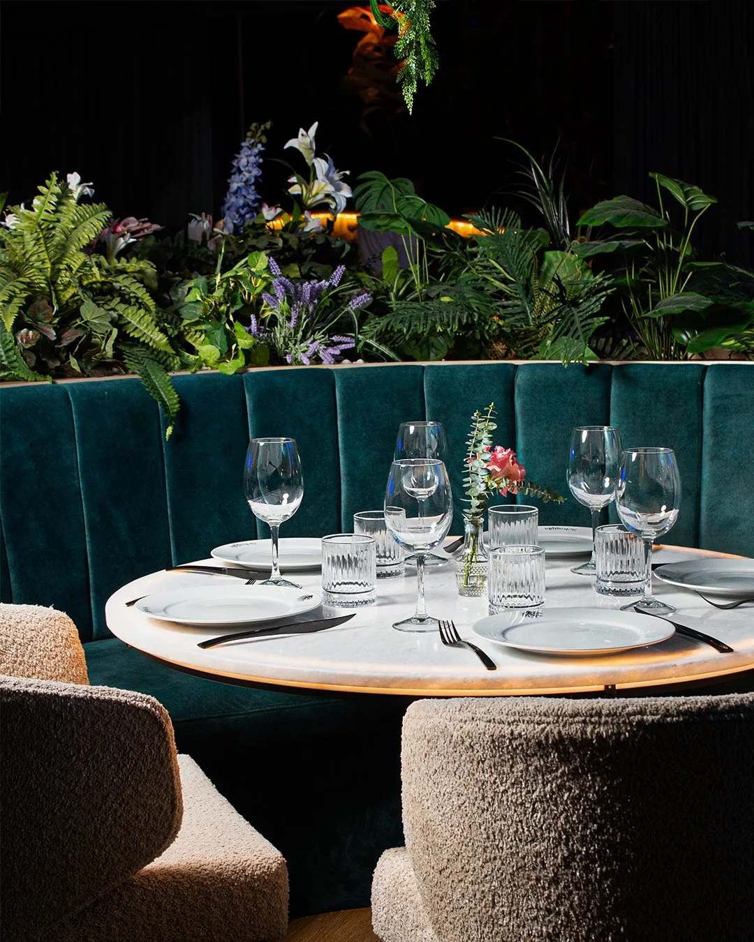 Restaurant table set with white plates, wine glasses, water glasses, silverware, and a small flower arrangement, surrounded by beige and teal seating, with lush green plants and flowers in the background.