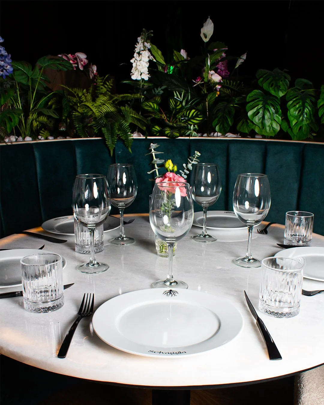 Table set for six with white plates, black utensils, and clear glasses, centerpiece with pink flowers, green plants background.