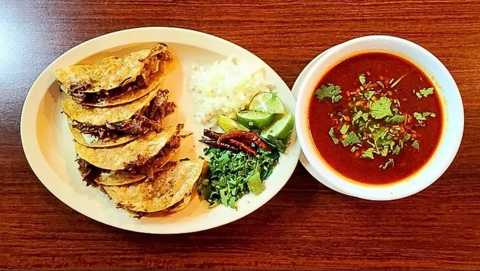 Plate of five slices of crispy beef, side of rice, lime wedges, cilantro, and chili peppers, with a bowl of spicy red soup garnished with cilantro.