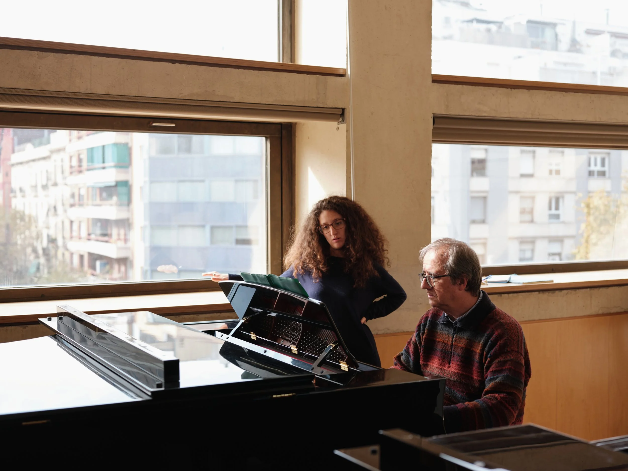 A young woman with curly hair and glasses sitting at a grand piano, with an older man sitting next to her, in a room with large windows overlooking city buildings.
