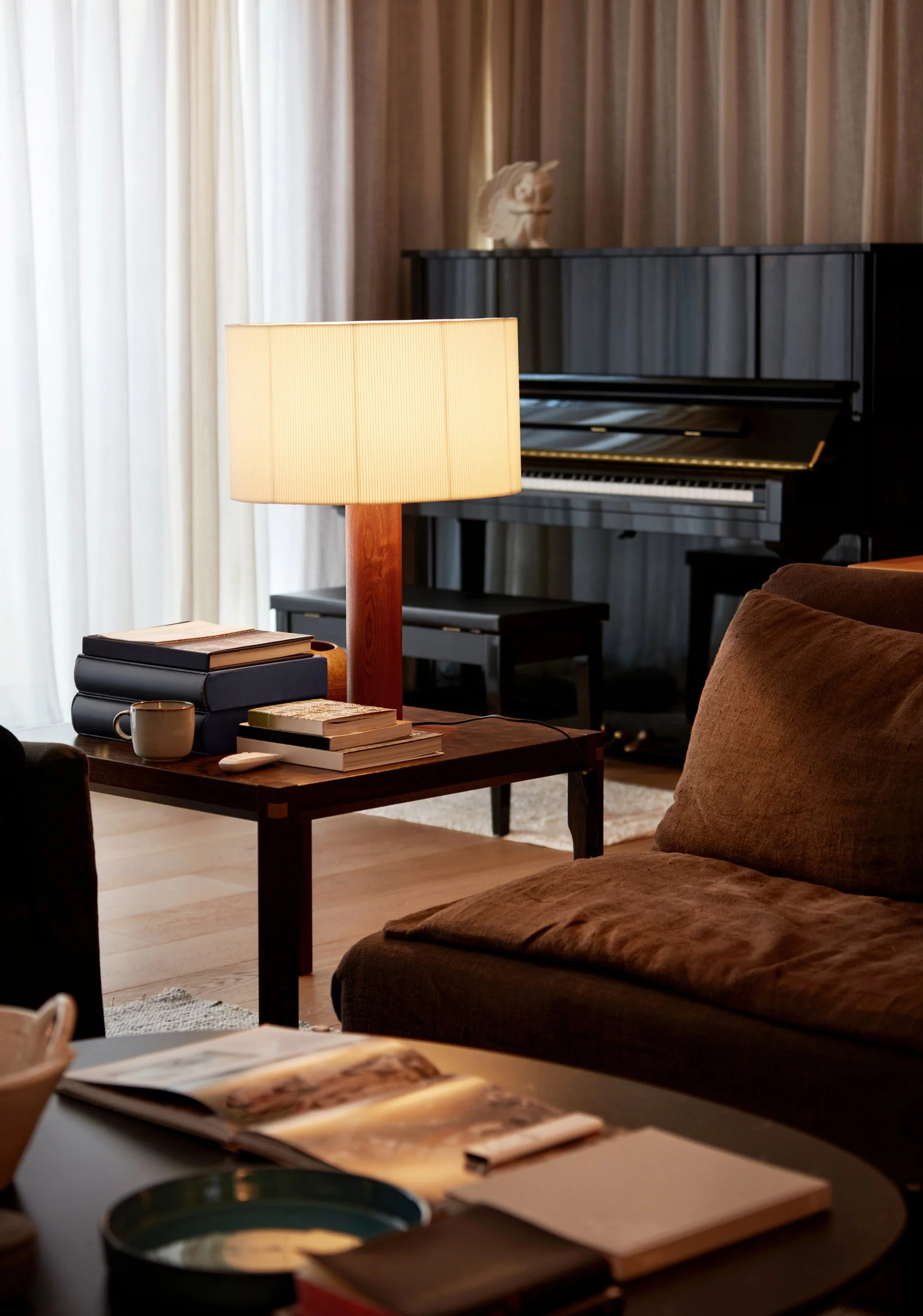 A cozy, dimly lit living room featuring a glowing wooden table lamp on a side table with books, with a black upright piano and a brown sofa in the background.
