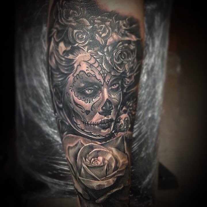 Tatouage noir et gris représentant une femme avec un visage maquillé comme un calavera mexicain, entourée de roses.