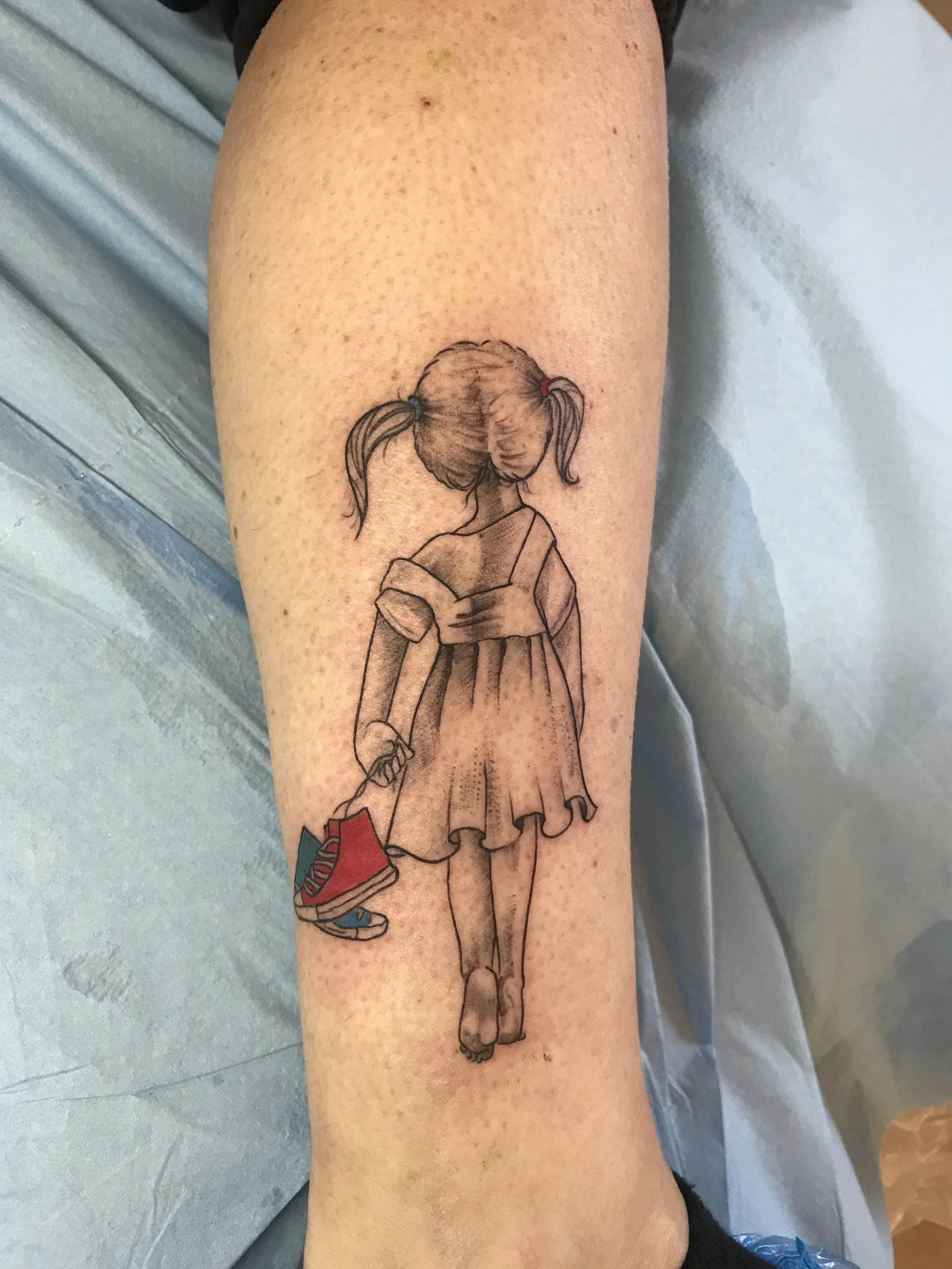 Tatouage d'une fille vue de dos, avec deux couettes, portant une robe et tenant une paire de chaussures de sport dans la main.