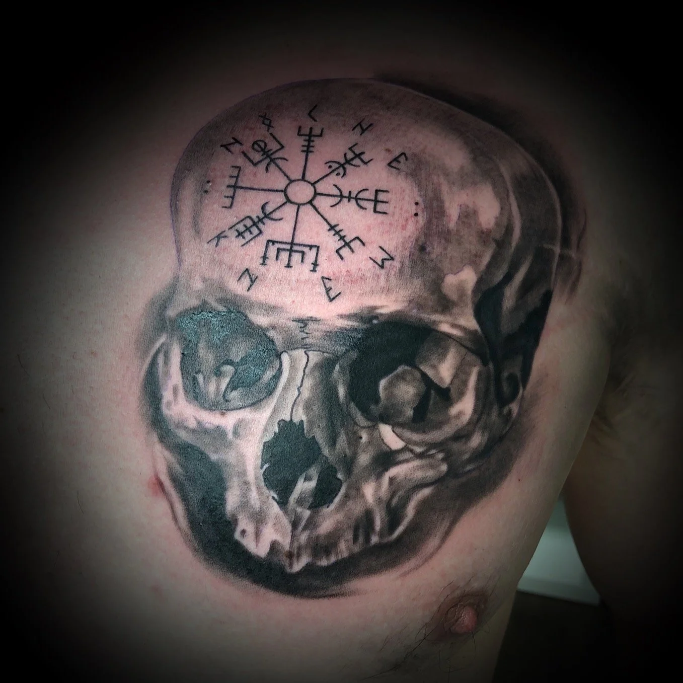 Tatouage d'un crâne humain en noir et gris avec un symbole nordique sur le haut du crâne.