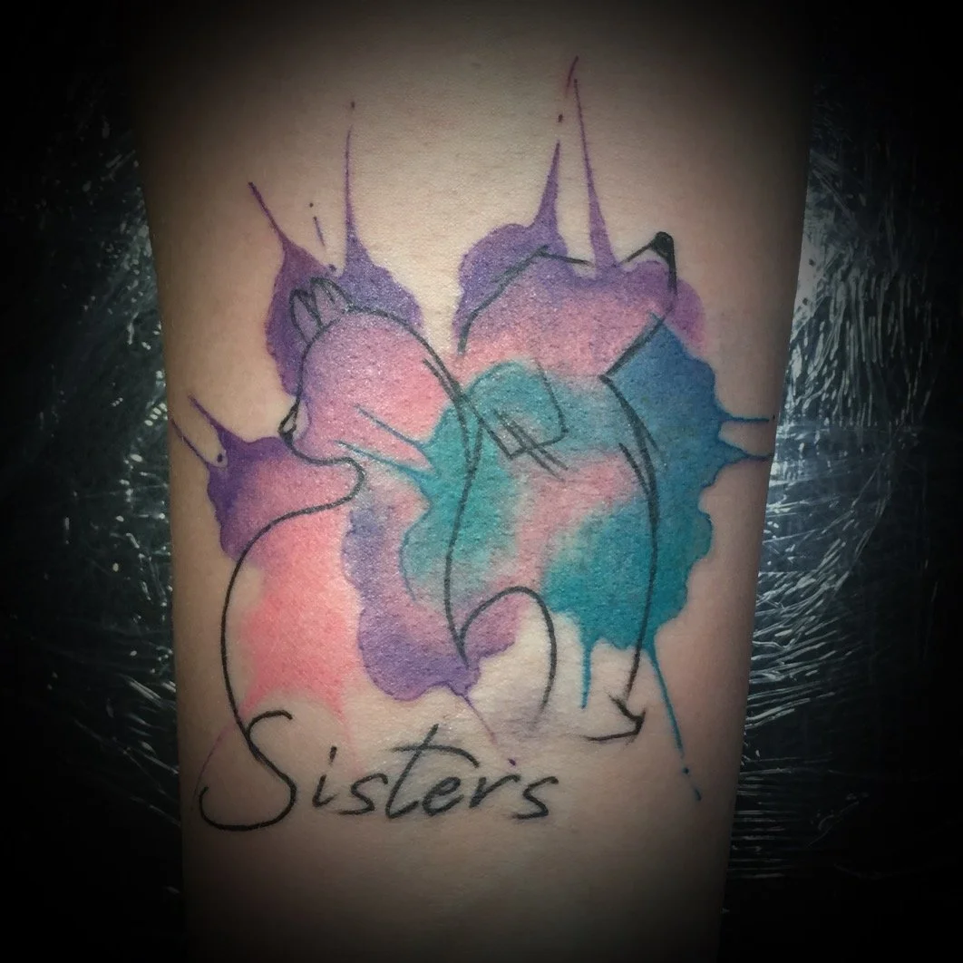 Tatouage aquarelle représentant deux visages de sœurs, avec la mention 'Sisters' en dessous.