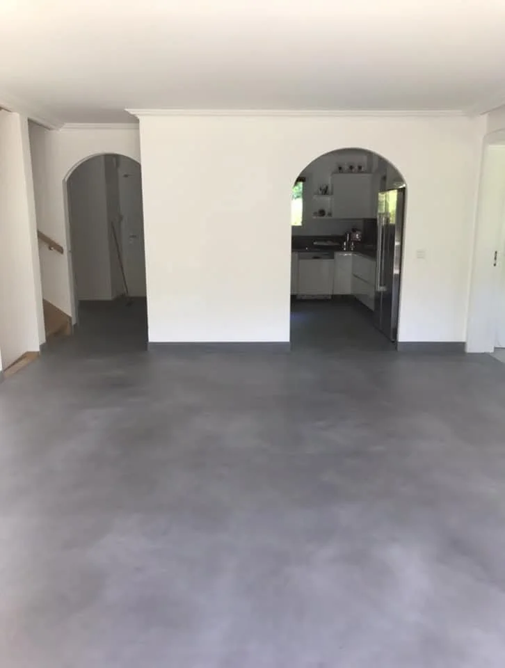 Intérieur d'une maison avec un mur blanc, un sol en béton gris, une cuisine visible à travers une arche, et un escalier en bois sur le côté gauche.