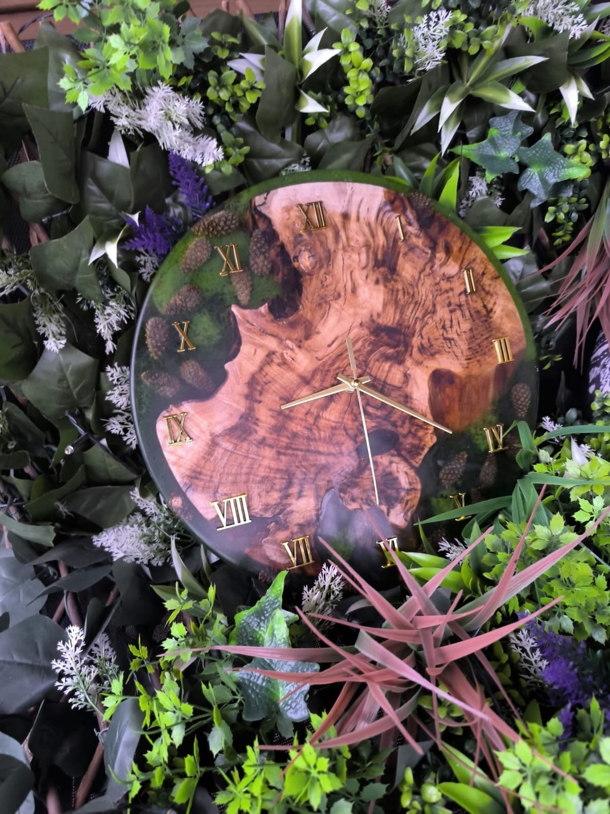 Horloge murale avec un fond en bois naturel et chiffres romains en or, entourée de nombreuses plantes vertes et fleurs.