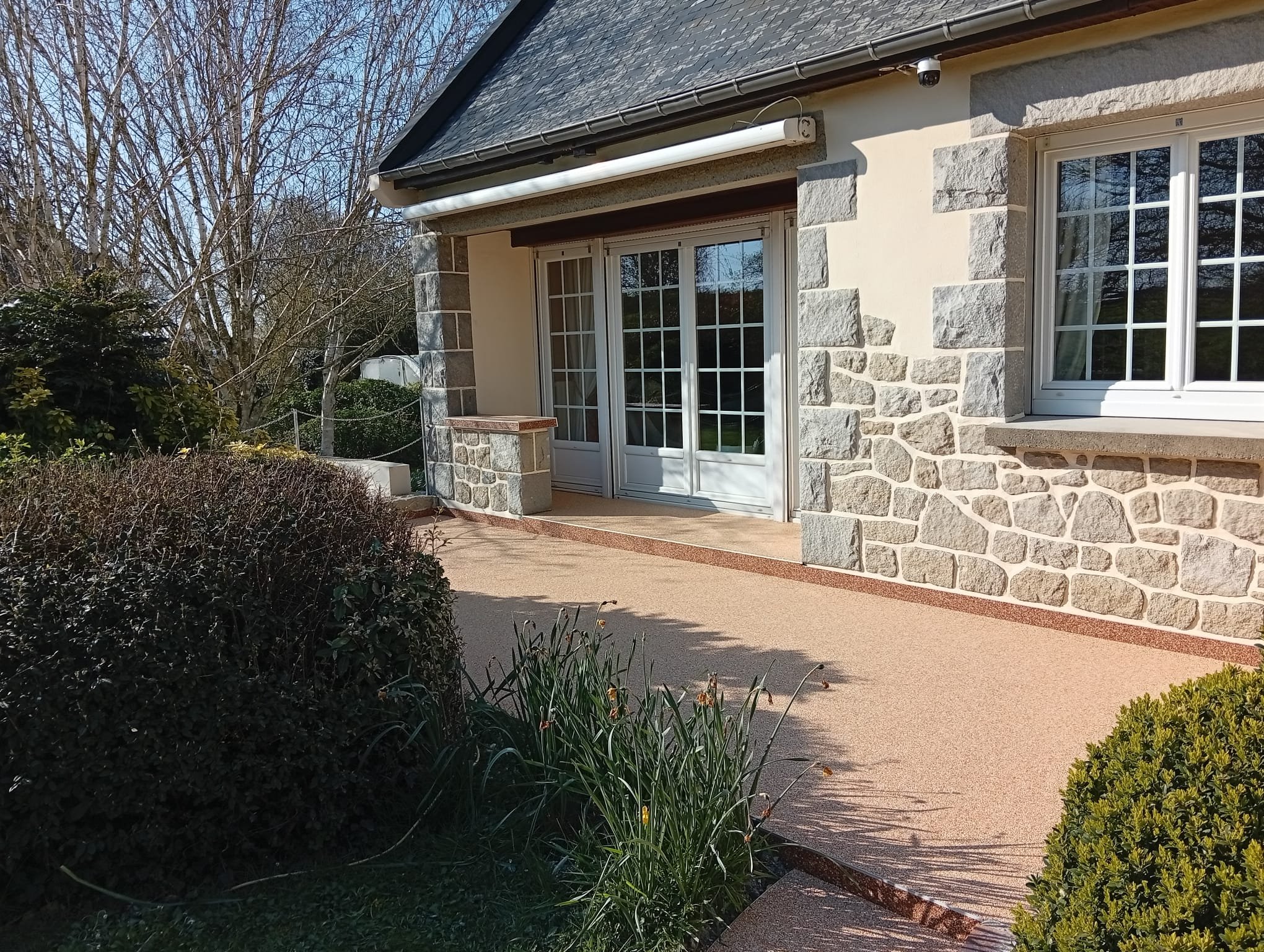 Façade d'une maison en pierre avec une terrasse en béton et des fenêtres à volets blancs