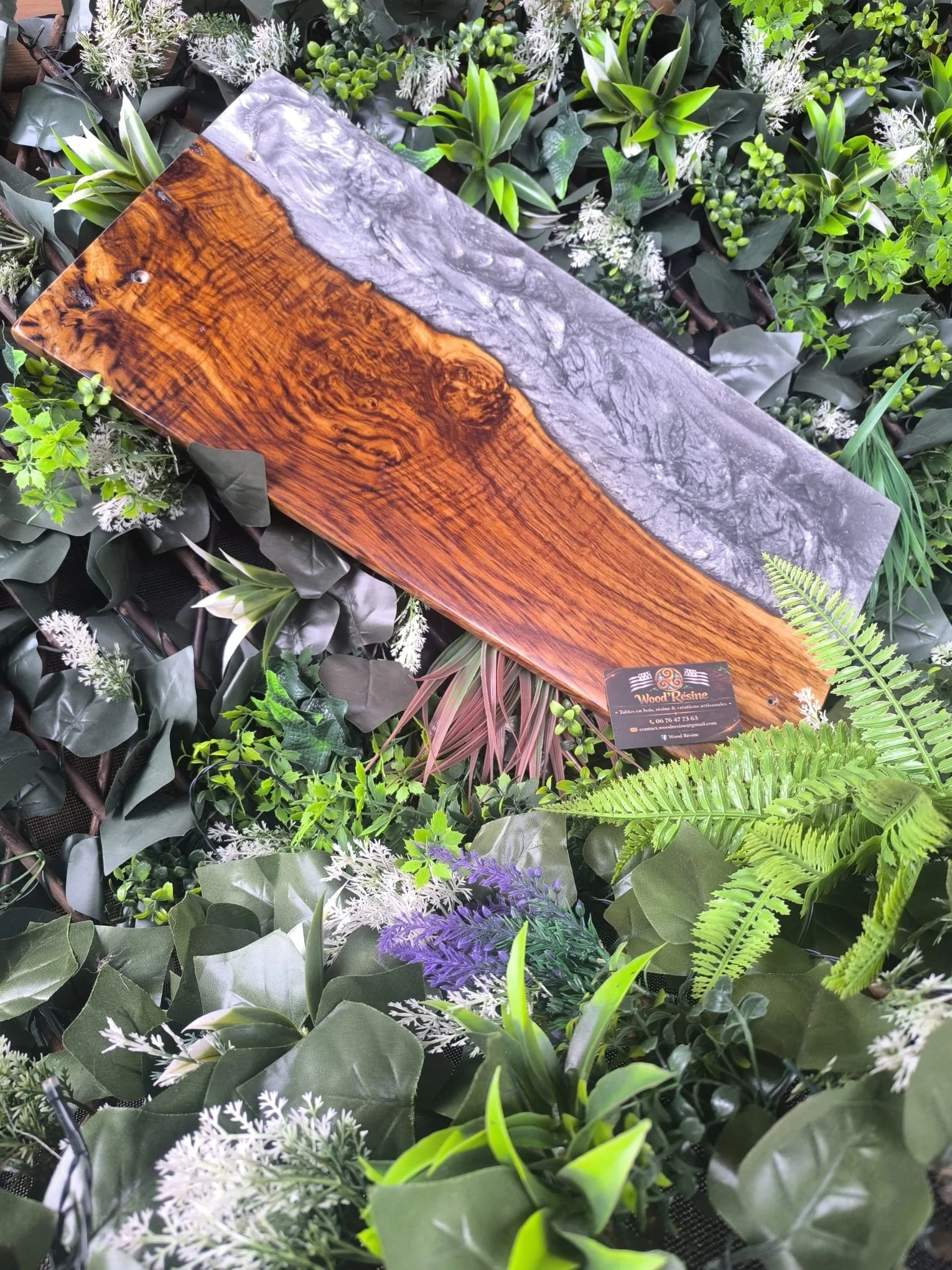 Une planche en bois mêlant bois poli et résine, posée sur un fond de plantes vertes et fleuries.