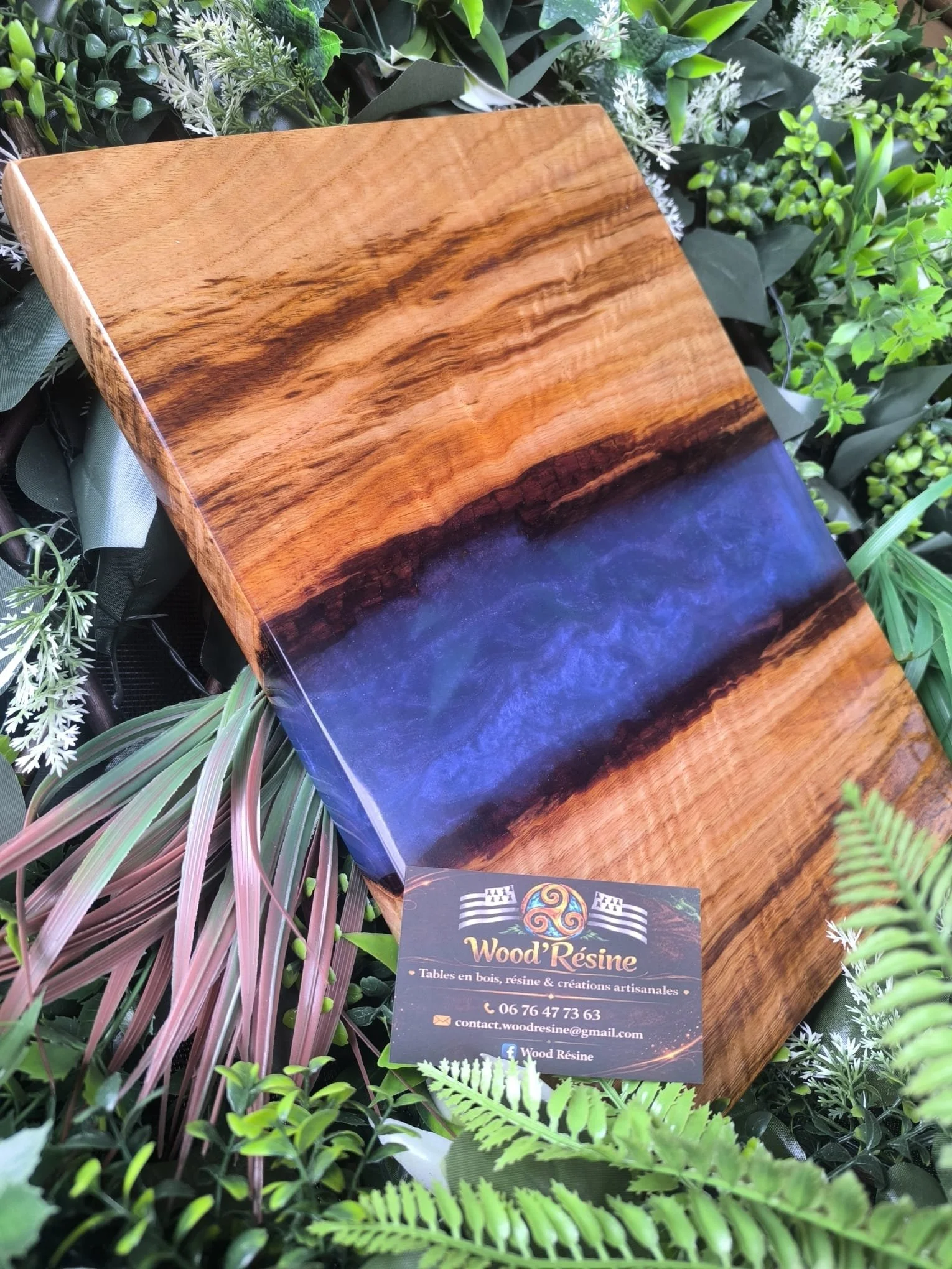 Planche en bois avec un insert en résine bleue, entourée de plantes vertes et une carte de visite pour 'Wood Résine'.