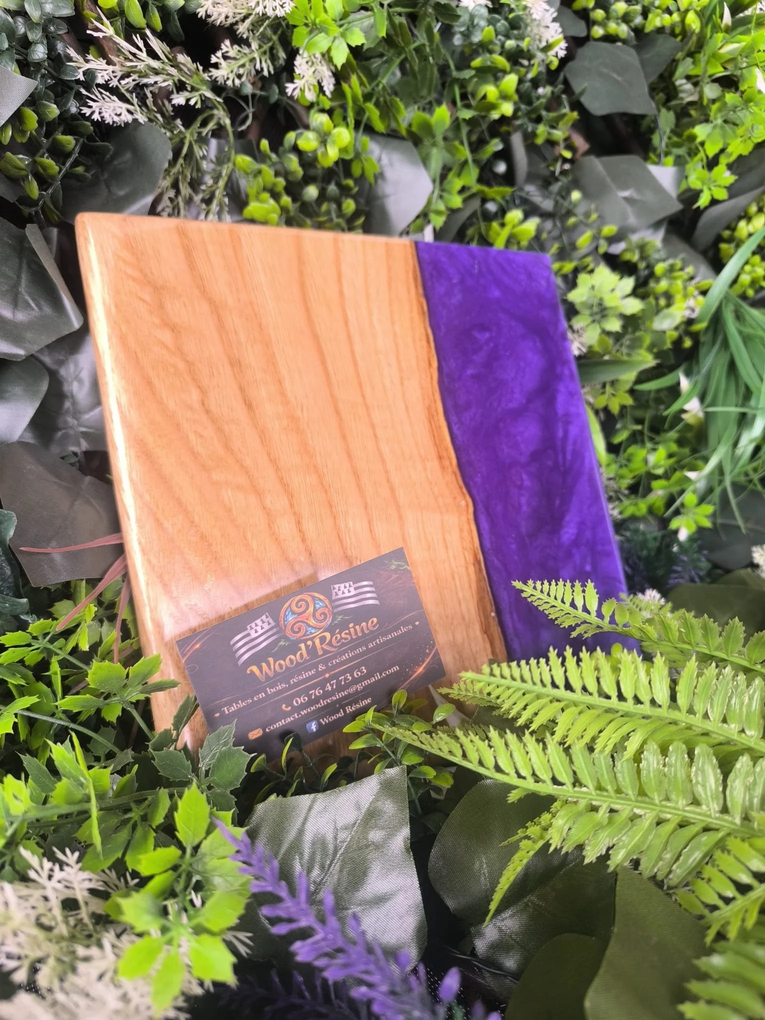 Planche en bois avec une section en résine violette, entourée de feuillage vert et de fleurs artificielles. Un petit panneau indique 'Wood Résine, Tables en bois, résine & créations artisanales'.
