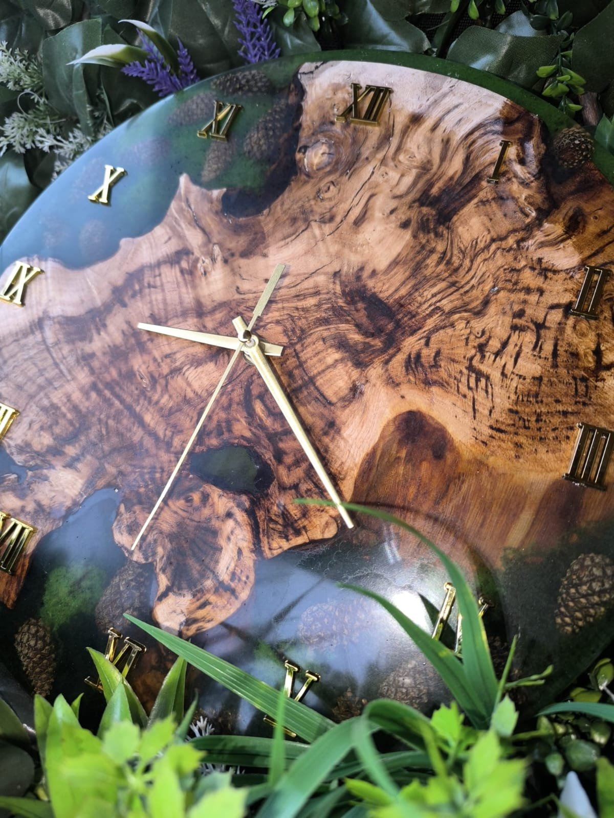 Horloge murale en bois avec chiffres romains dorés, entourée de feuillage vert.