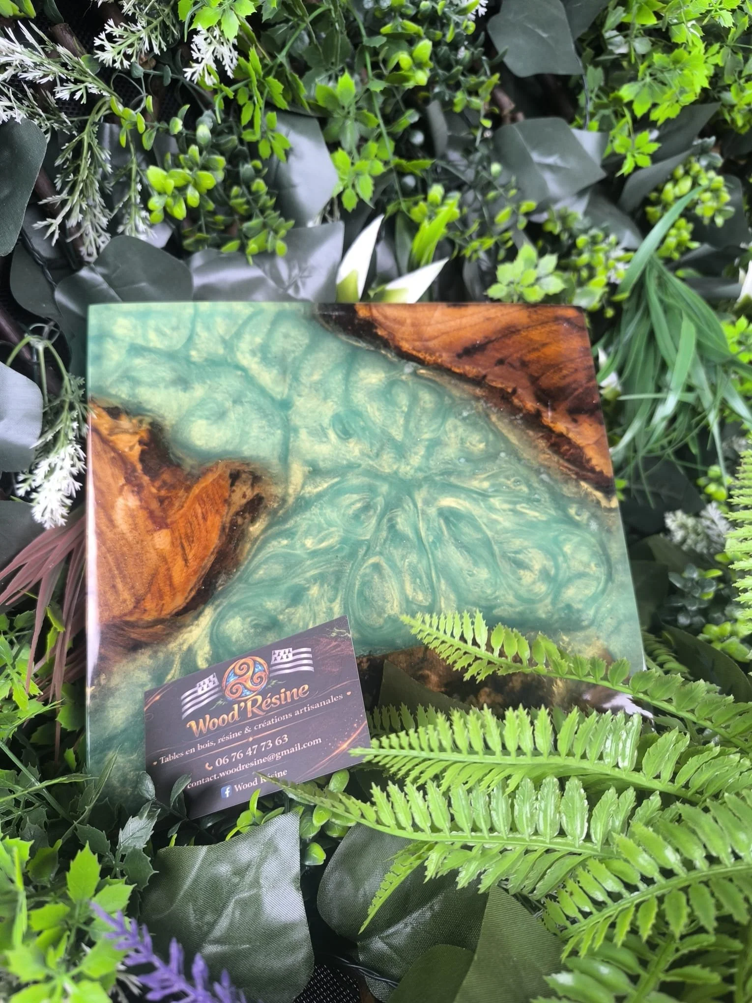 Une tablette en résine de bois, verts et marron, posée parmi des feuillages verts variés.