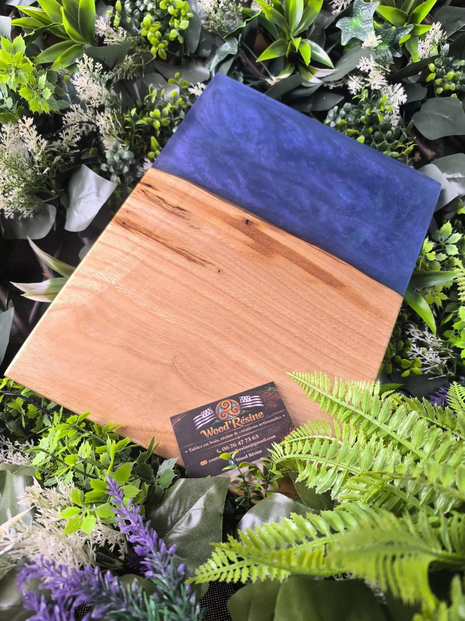 Une planche en bois avec une partie recouverte de résine bleue, entourée de diverses plantes vertes et d'une carte de visite de 'Wood Résine'