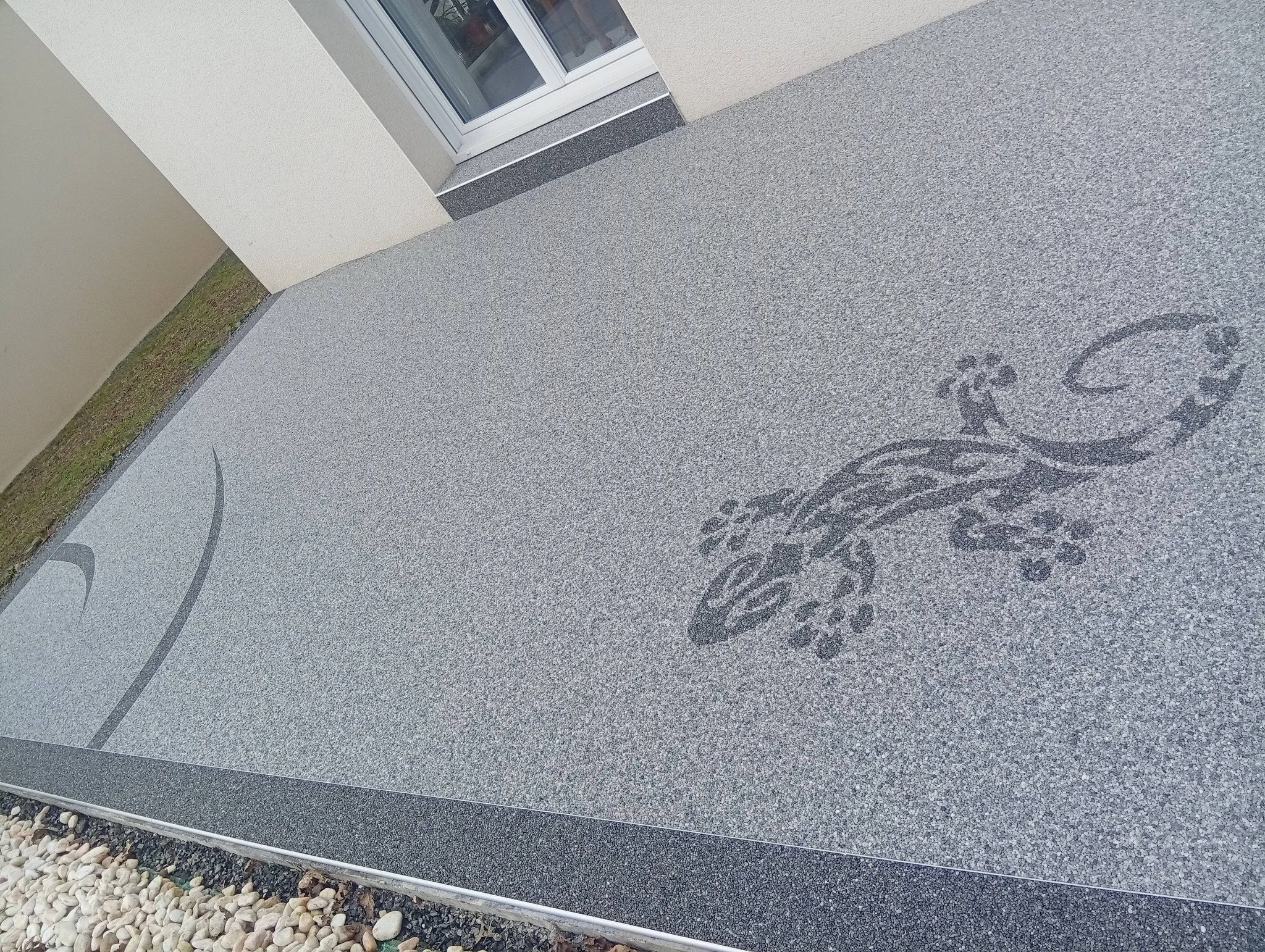 Terrasse en moquette de pierre avec une mosaïque de lézard noir en bas à droite, façade d'une maison avec porte fenêtre en arrière-plan.