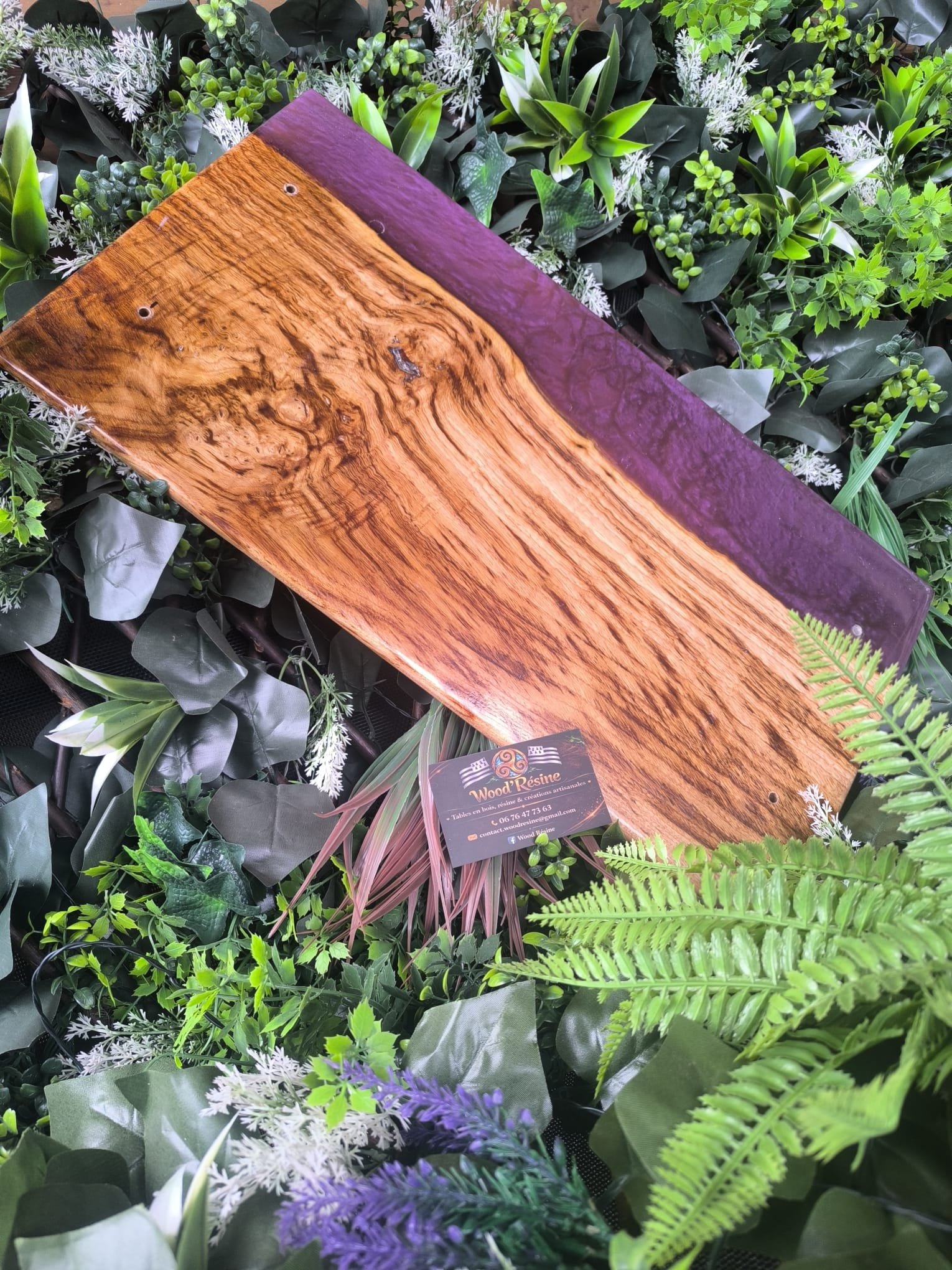 Planche en bois avec une bordure violette, entourée de plantes vertes et de fougères.