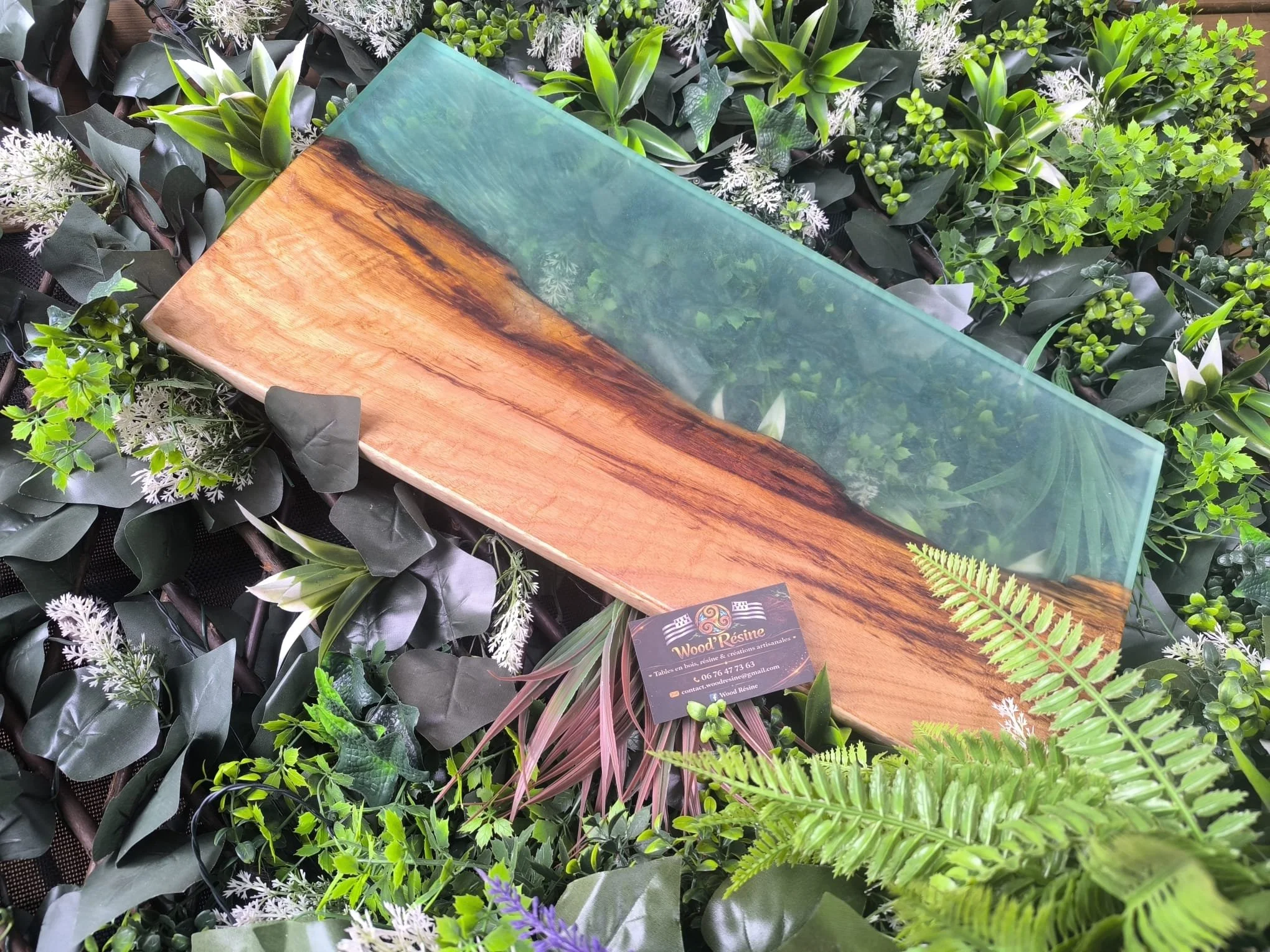 Table en bois avec un insert en résine transparente, posé sur un tapis de plantes vertes, dont des fougères, des feuilles de vin, et des fleurs blanches et violettes.