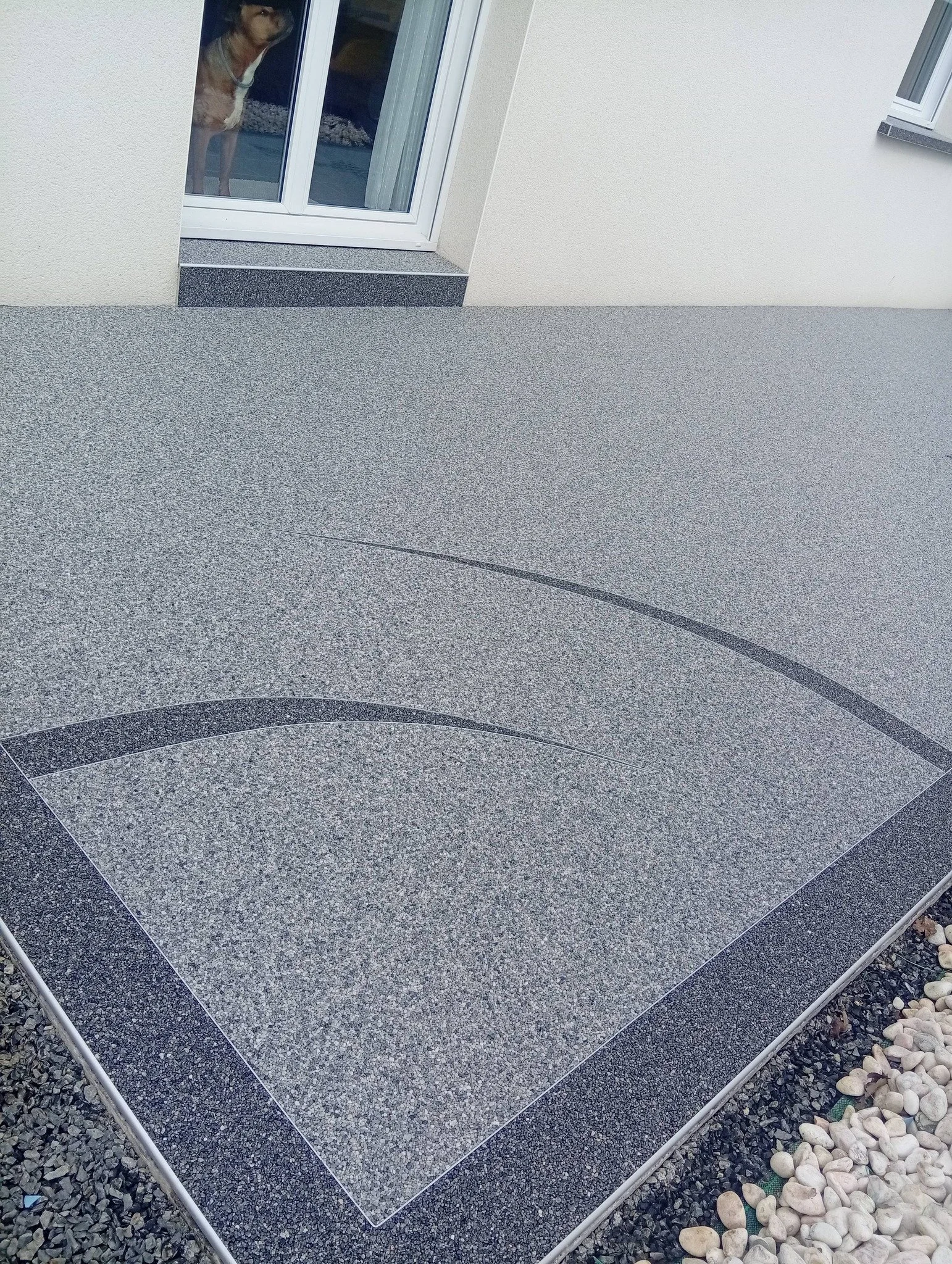 Terrasse en moquette de pierre avec un motif de lignes courbes, à côté de granulat de marbre gris clair et gris obscur, en face d'une porte-fenêtre dans un bâtiment blanc.