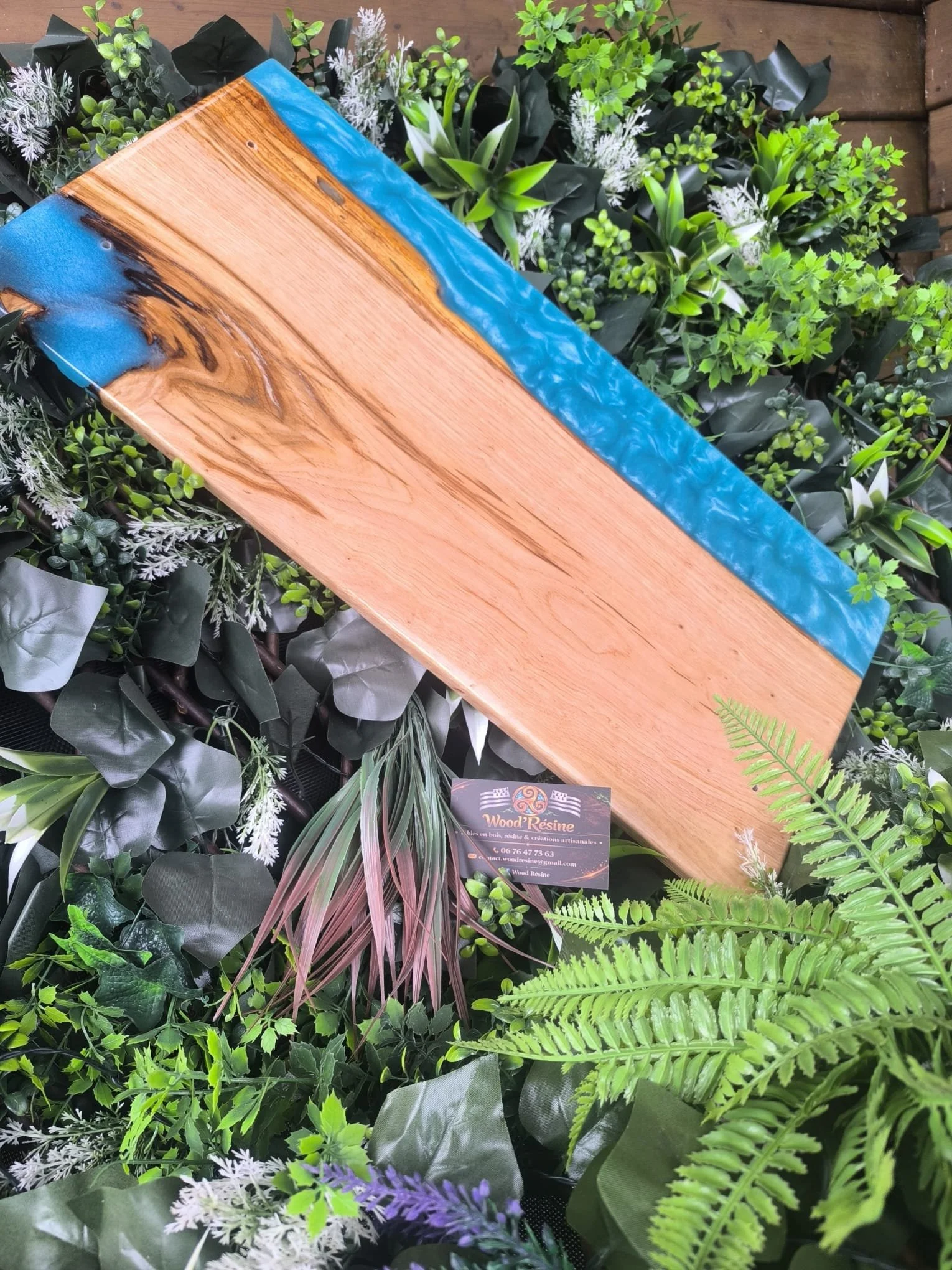 Une planche en bois avec une bordure en résine bleue, posée sur un tapis de plantes vertes variées, dont des fougères, des feuilles d'aspidistra et des feuilles à motifs violets et roses.