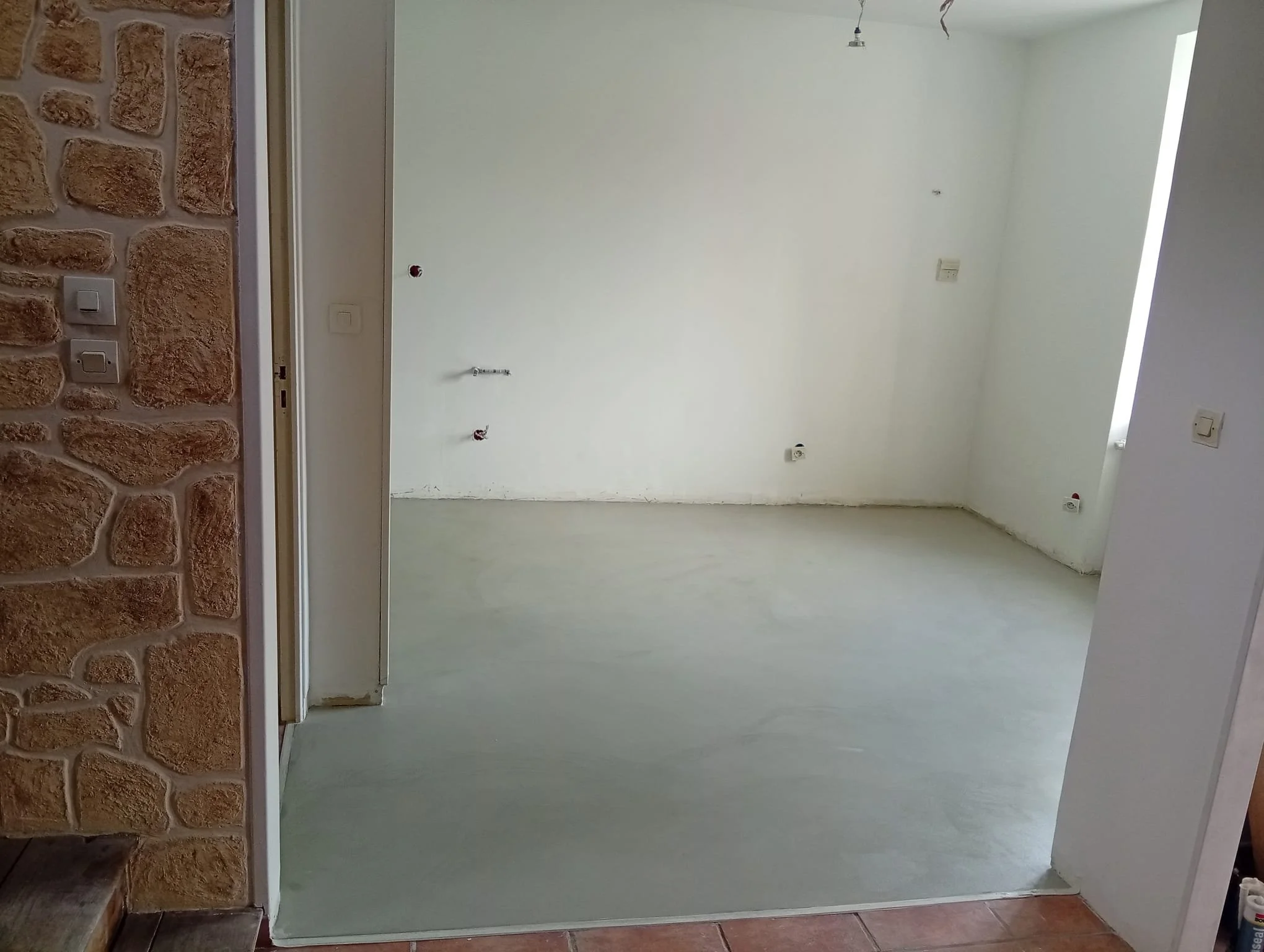 Intérieur d'une pièce en cours de rénovation avec murs blancs, sol en béton lissé, et plusieurs prises électriques sans plaques en place.