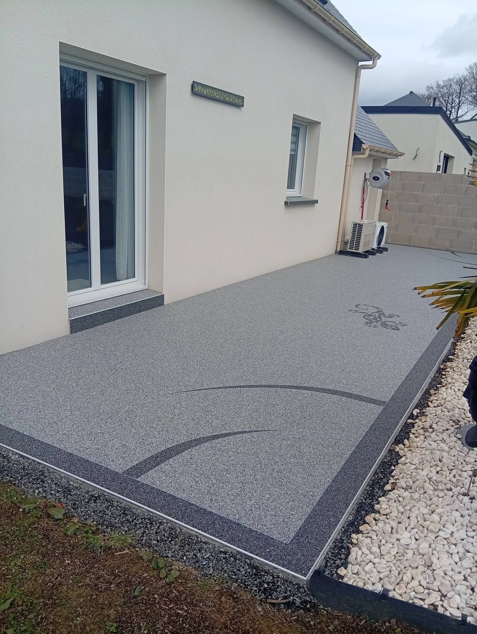 Une terrasse en moquette de pierre attenante à une maison moderne avec un mur blanc, des fenêtres et une porte-fenêtre laissant entrer la lumière. La terrasse est décorée avec un motif artistique et est bordée de petites plantes.