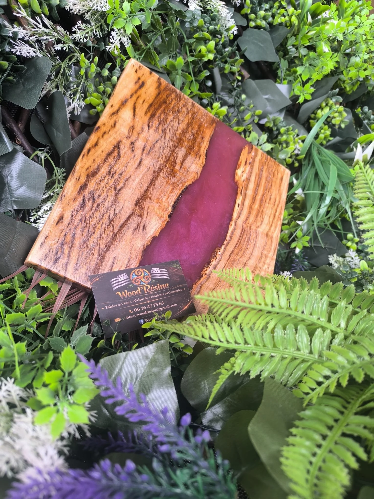 Une plaque en bois et résine de couleur violette entourée de plantes vertes et de feuillages.