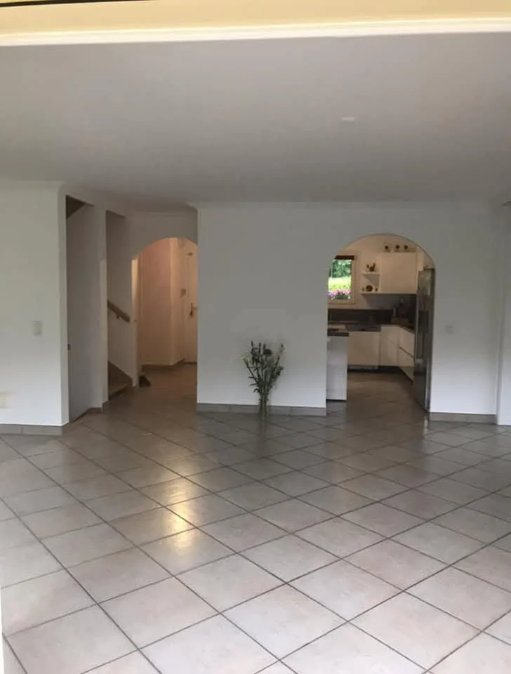 Intérieur d'une maison avec sol en carrelage, mur blanc, et porteurs d'arcades menant à la cuisine et à une autre pièce, avec un vase contenant des fleurs au centre.
