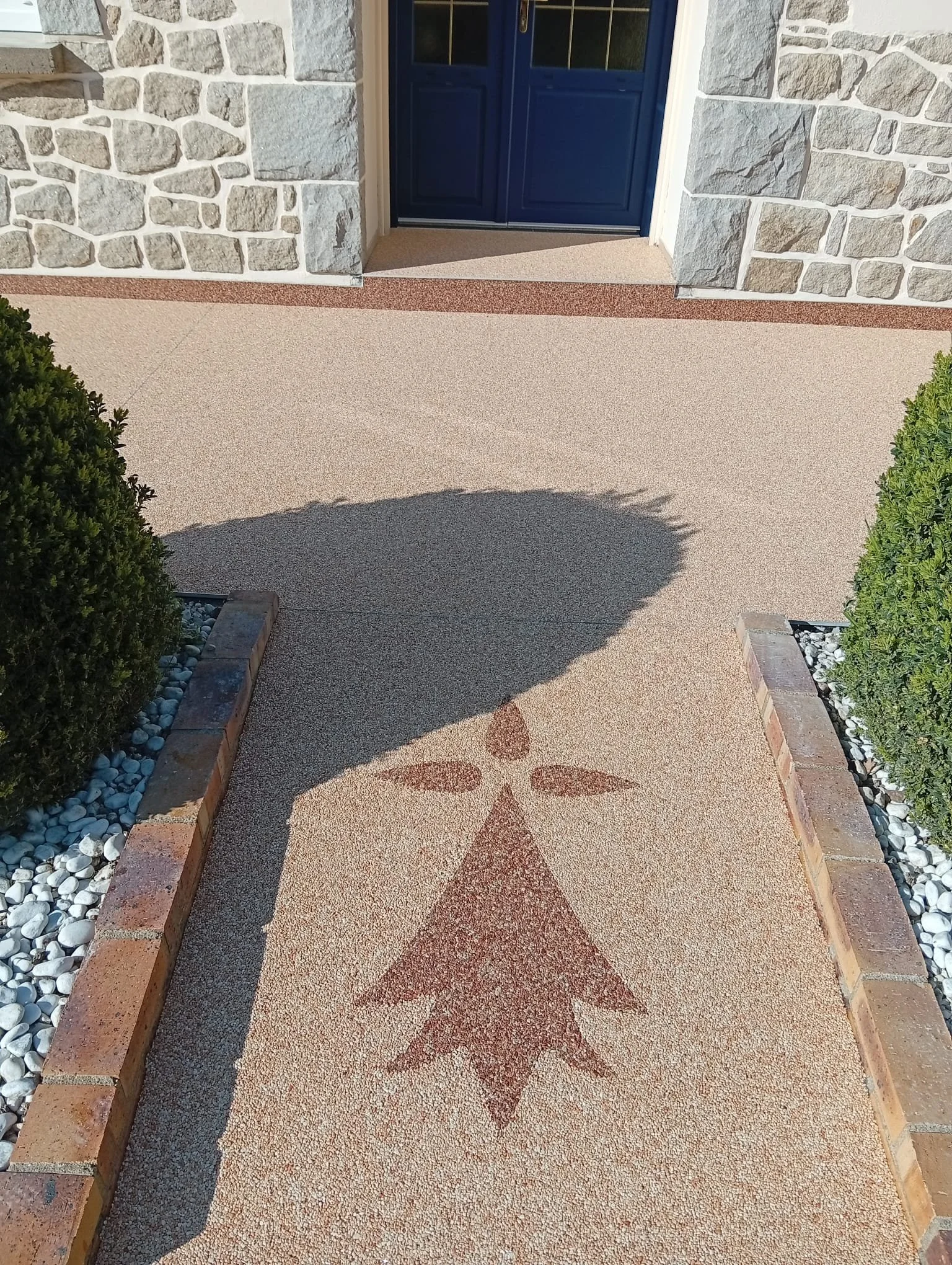 Chemin en béton décoratif avec un motif d'un arbre à l'entrée d'une maison, entouré de bordures en briques et de buissons.